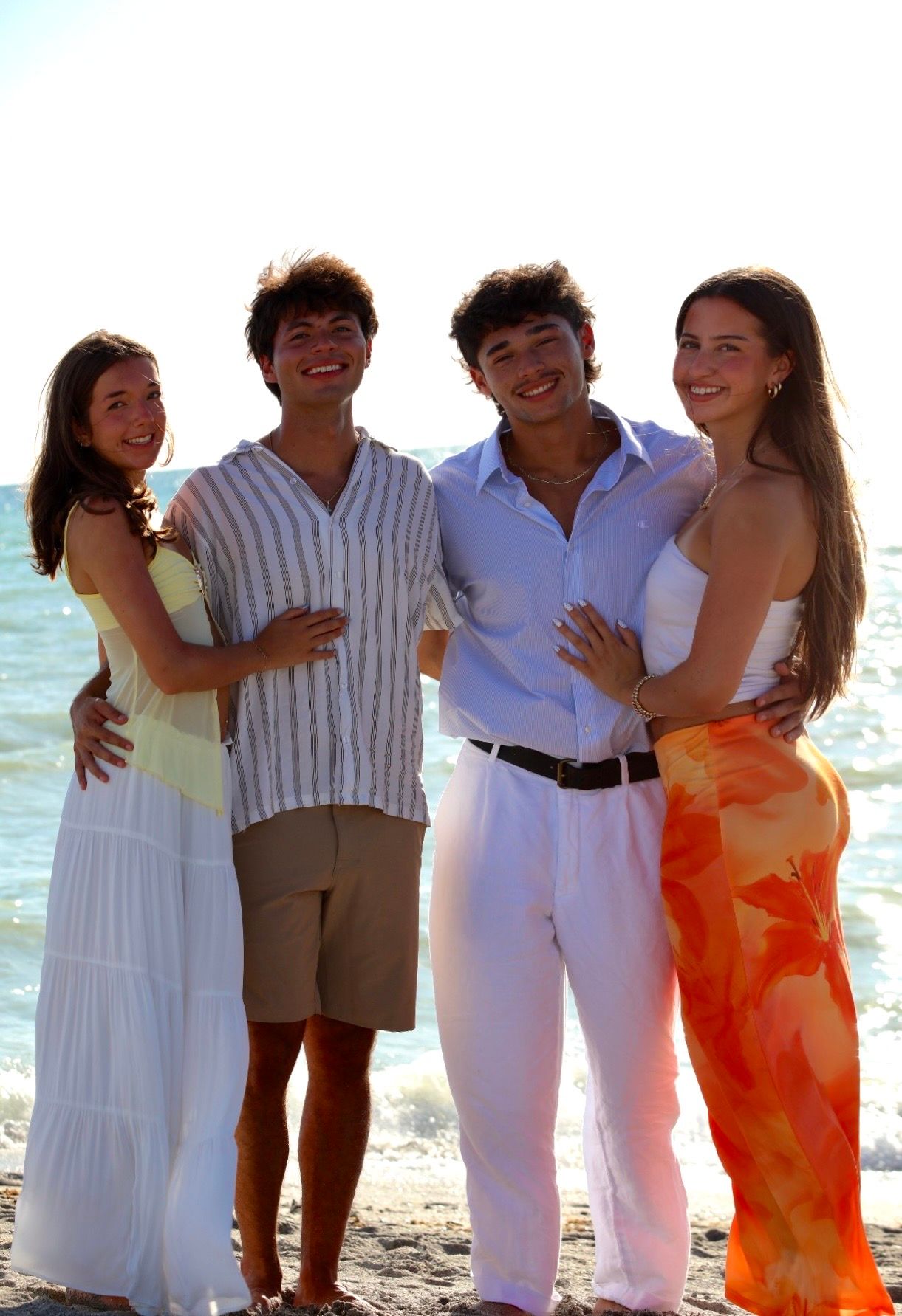 Two young couples posing together on a sunny beach, smiling in colorful summer outfits.