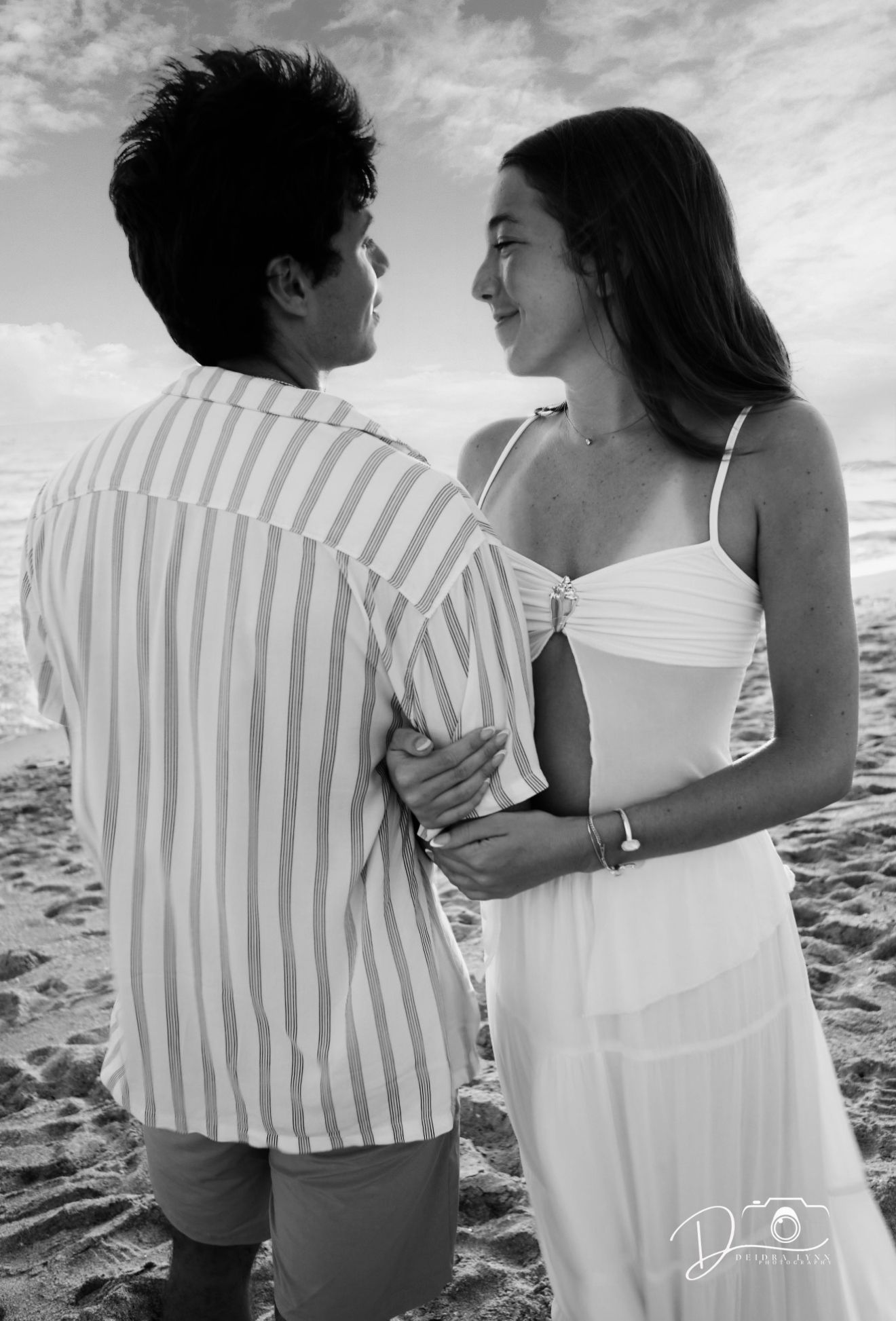 Black and white photo of couple embracing on beach, woman in white dress and man in striped shirt gazing at each other.