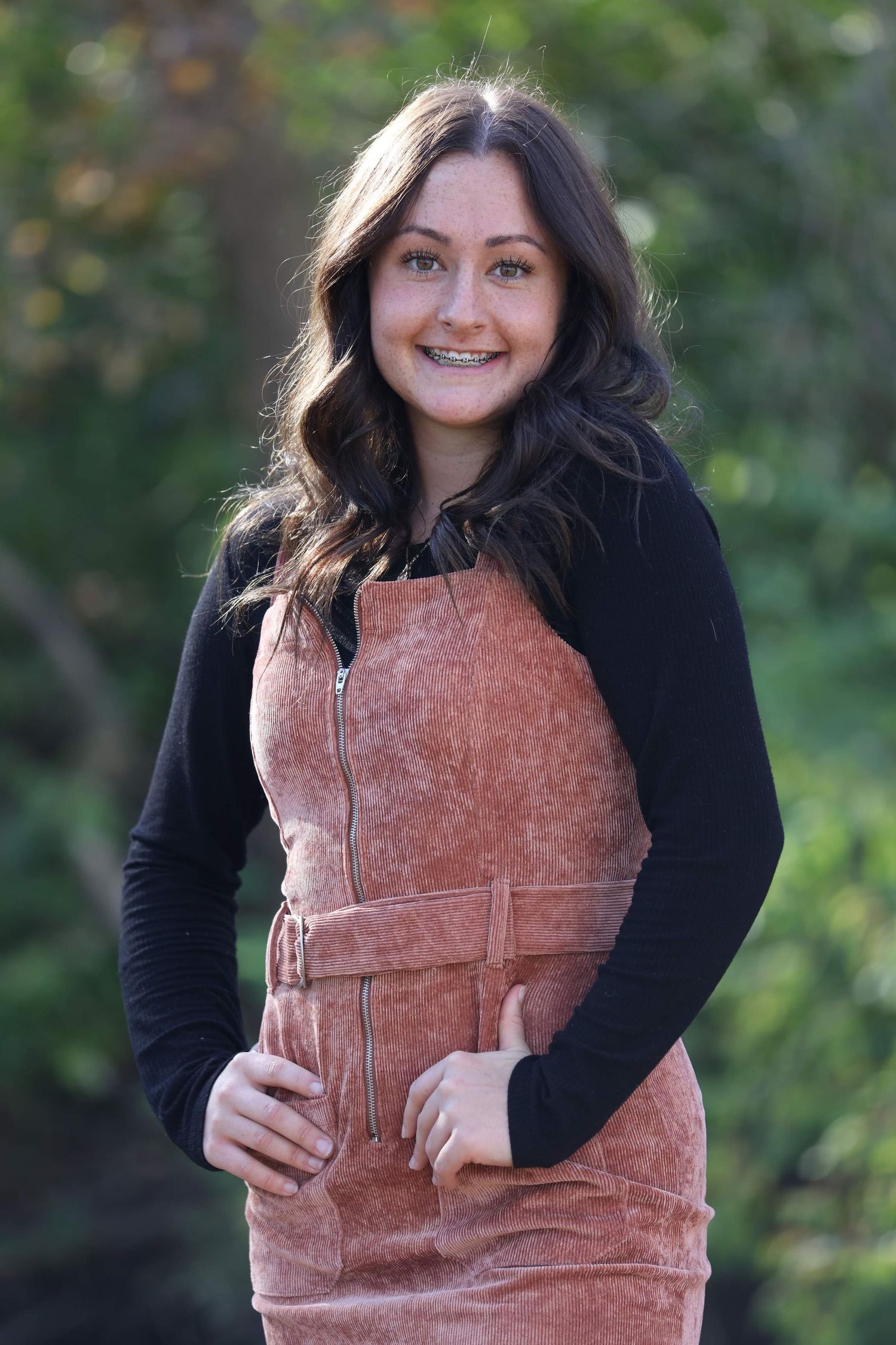 Smiling teen girl with braces wearing rust corduroy overalls over black long sleeve shirt outdoors.