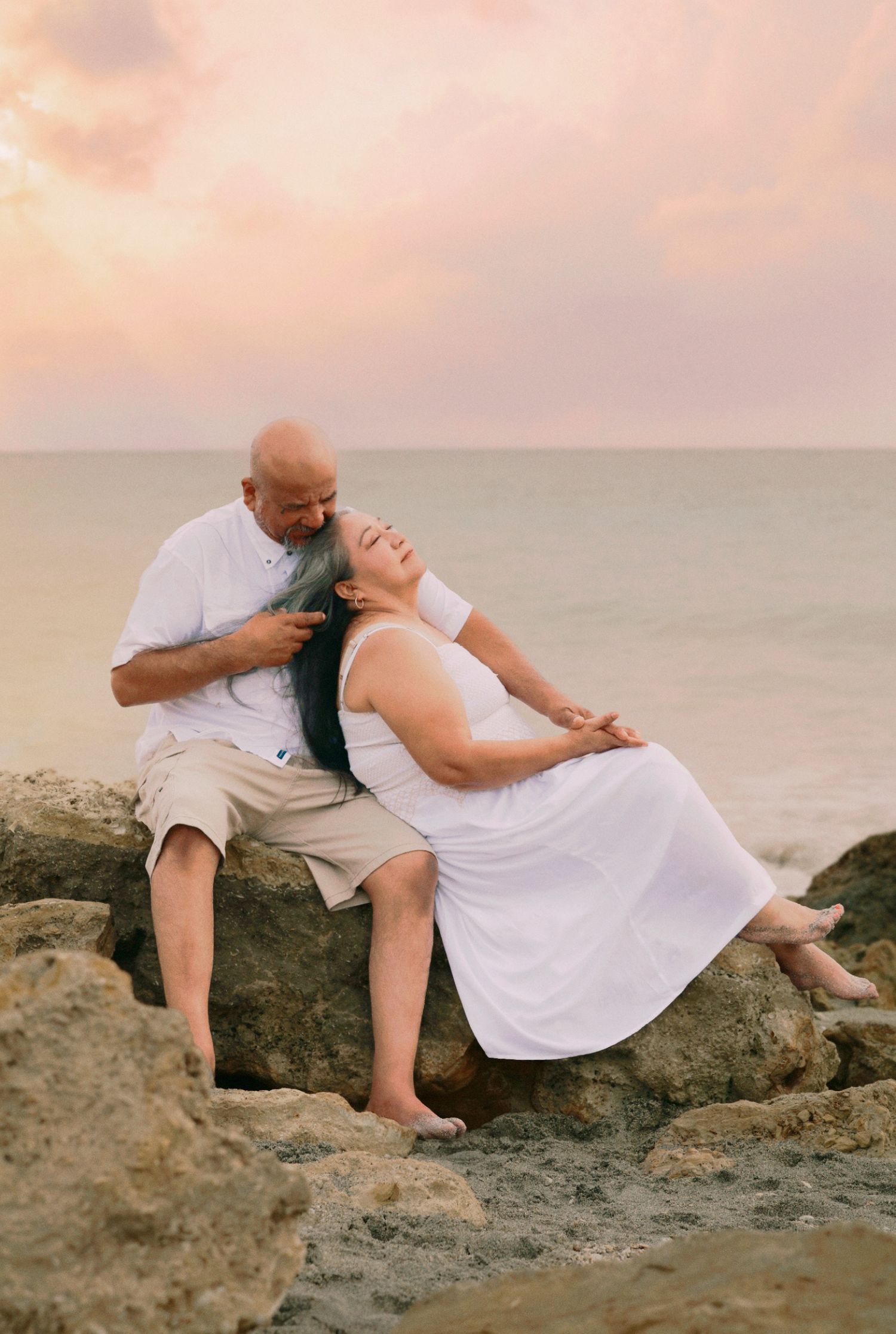 Romantic couple sitting on rocks by ocean at sunset, woman with teal hair leaning back.