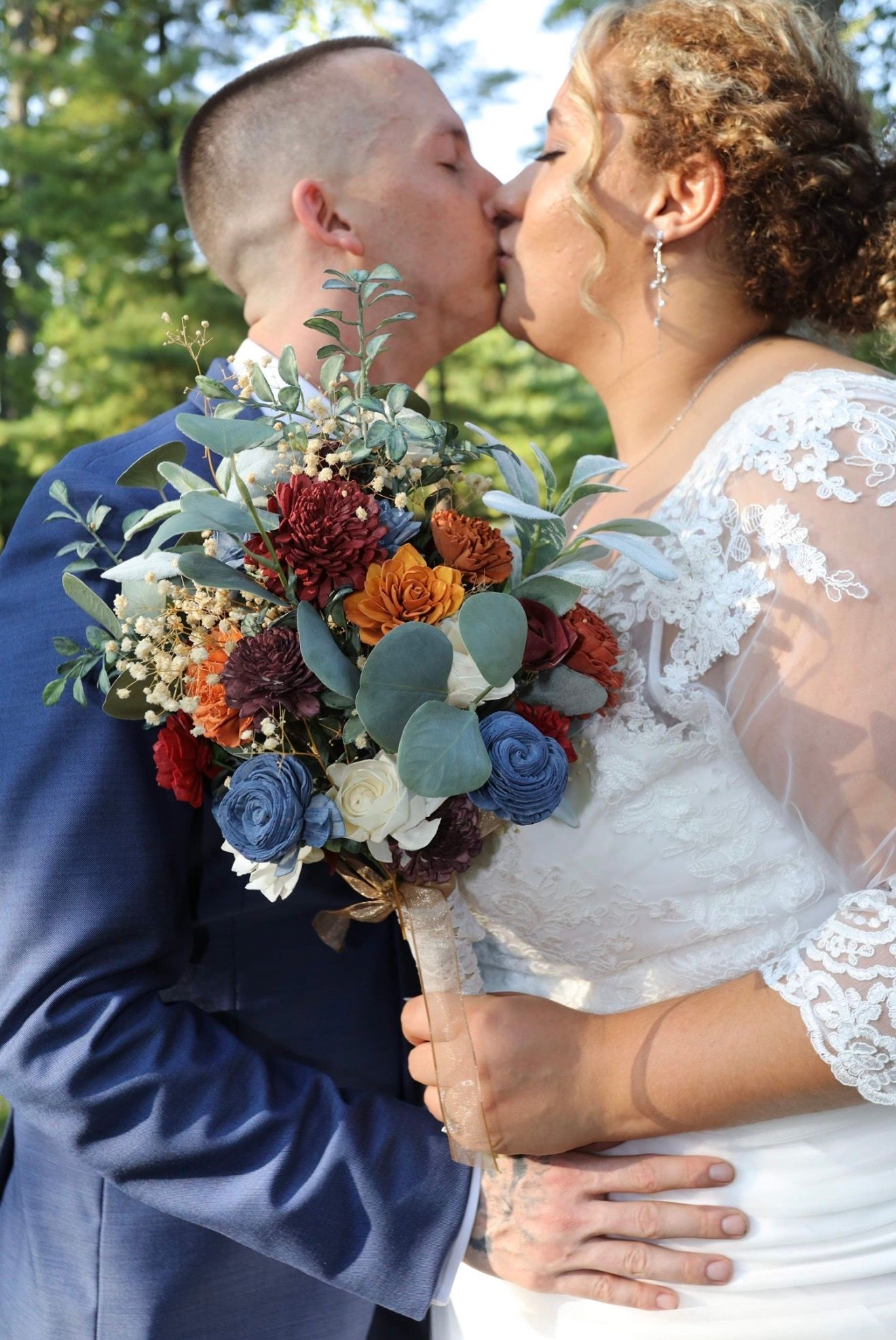 Bride and groom sharing a kiss, bride holding a bouquet of blue, burgundy, and orange flowers, outdoors.