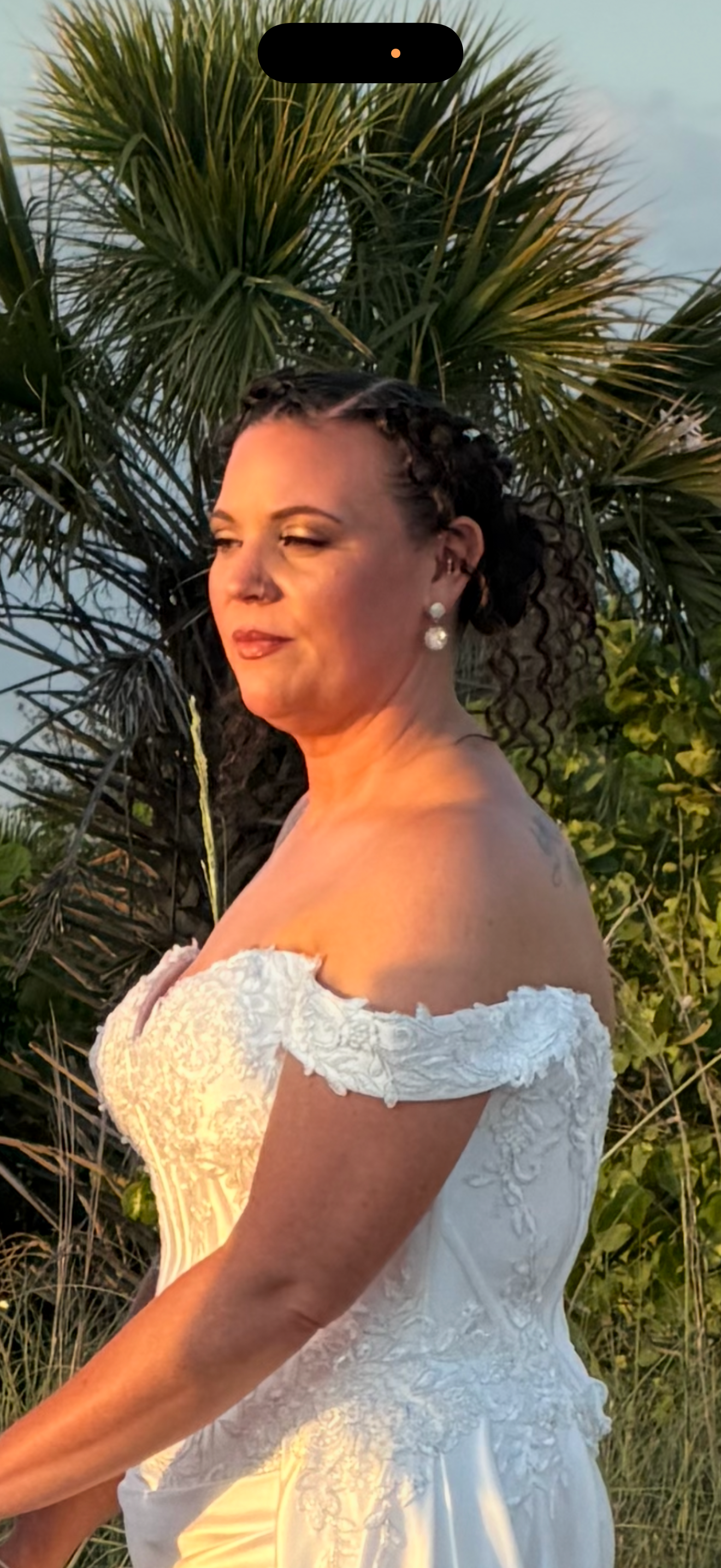 Bride in off-shoulder white lace wedding gown with pearl earrings and updo hairstyle, posing outdoors near palm trees.