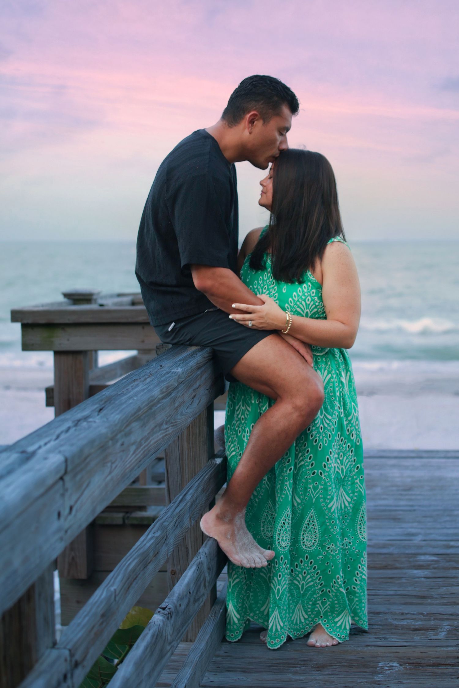Couple sharing a romantic moment on a wooden beach boardwalk at sunset, woman in green maxi dress.
