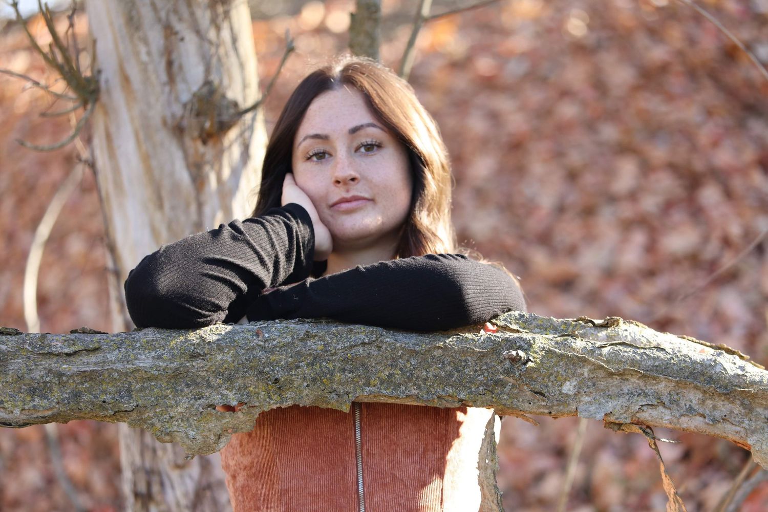 Young brunette woman in black gloves resting arms on a mossy log in an autumn forest setting.