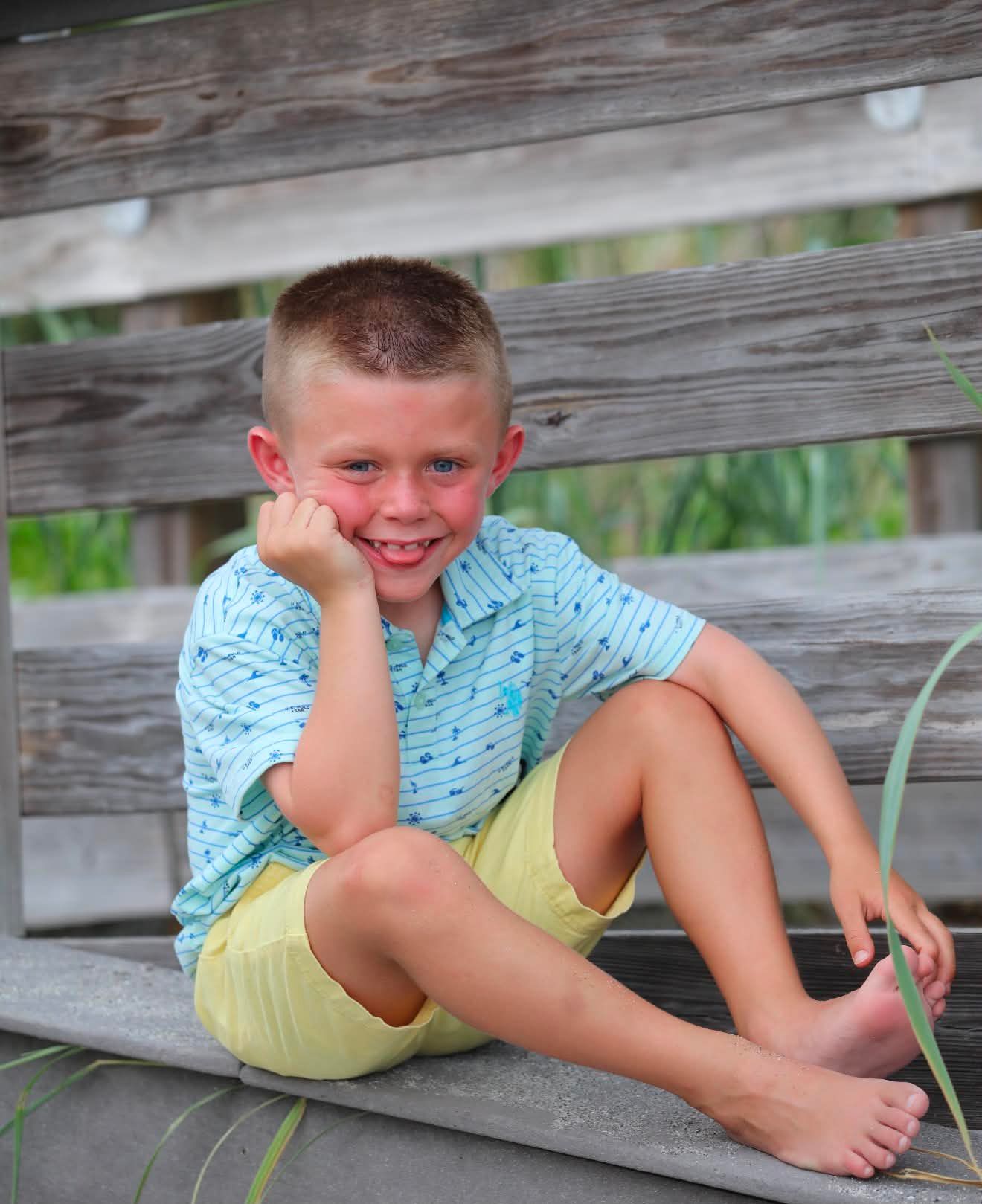 Smiling young boy in blue shirt and yellow shorts sitting barefoot on wooden steps outdoors.