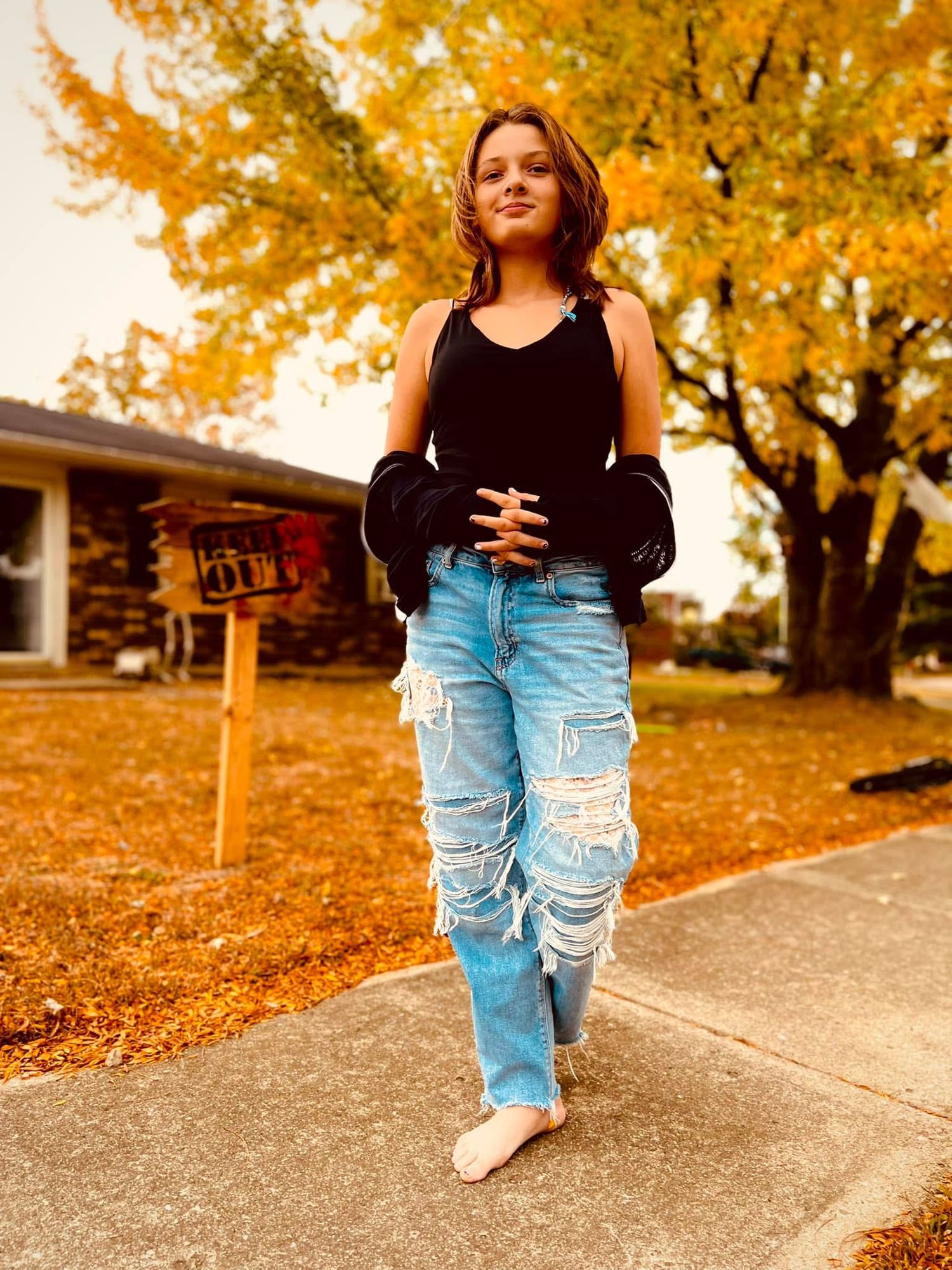 Young woman in ripped jeans and black top poses barefoot on sidewalk surrounded by autumn foliage.