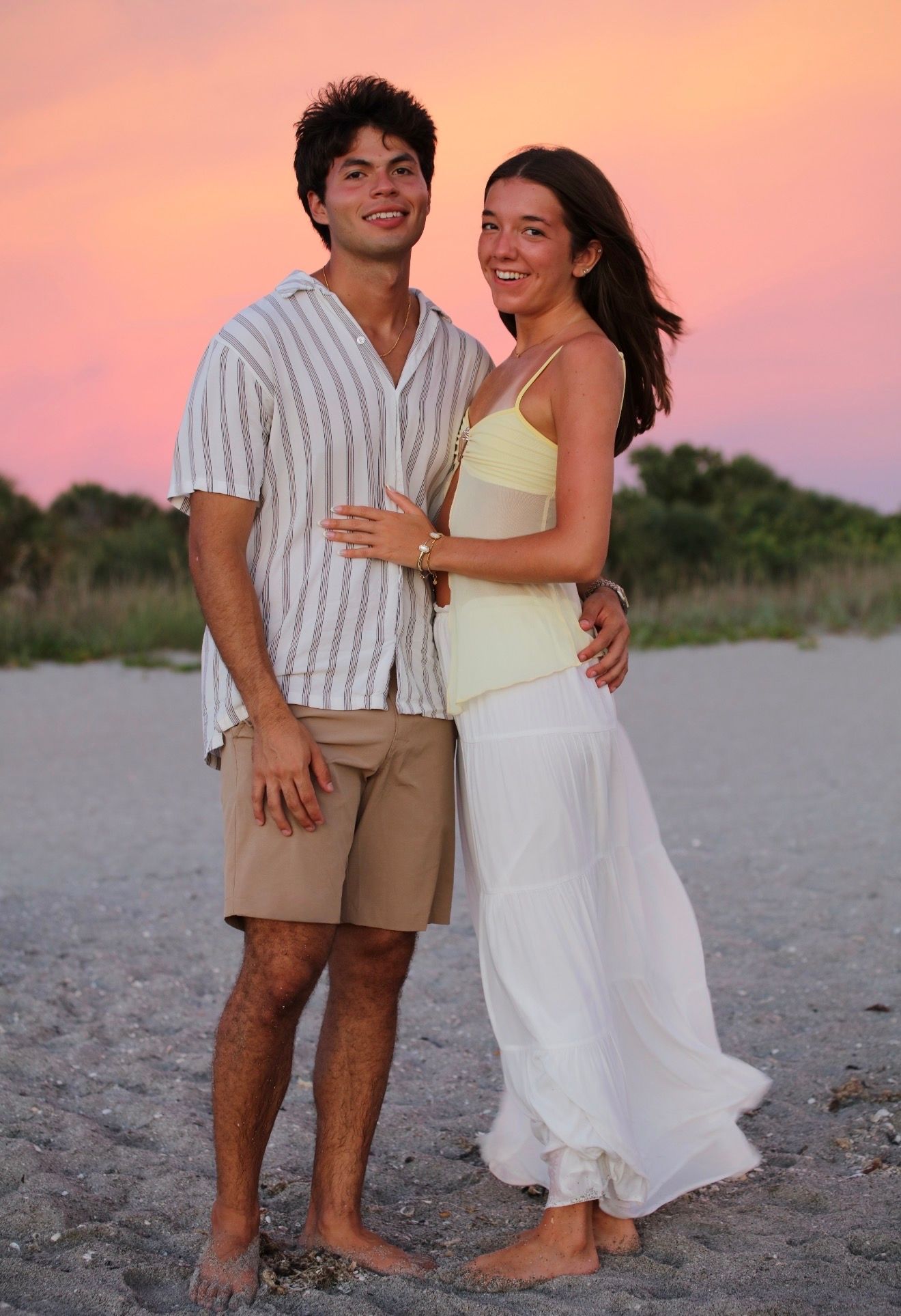 A smiling couple embraces on a sandy beach during a vibrant pink and orange sunset.