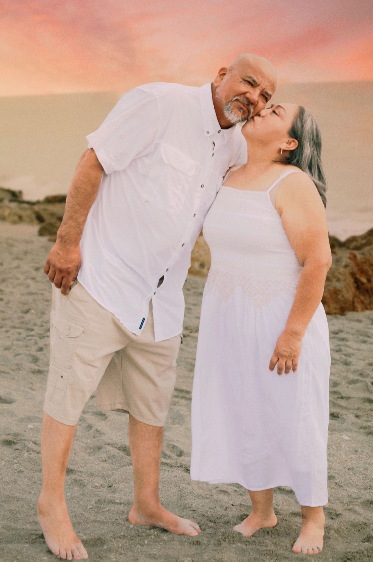 Couple in matching white outfits sharing a romantic kiss on a sandy beach at sunset.