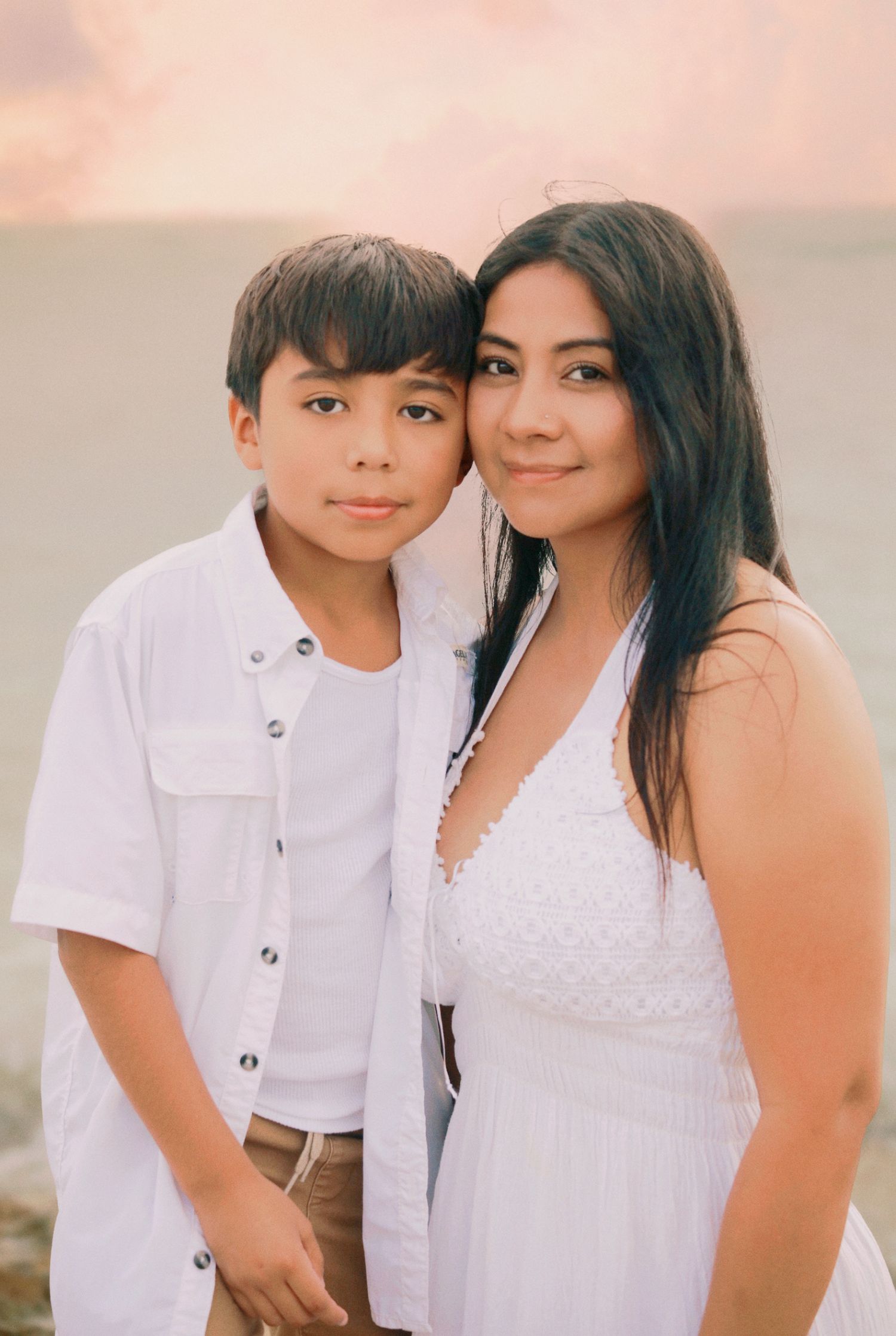 A young boy and woman in white outfits pose together at a beach during a colorful sunset.