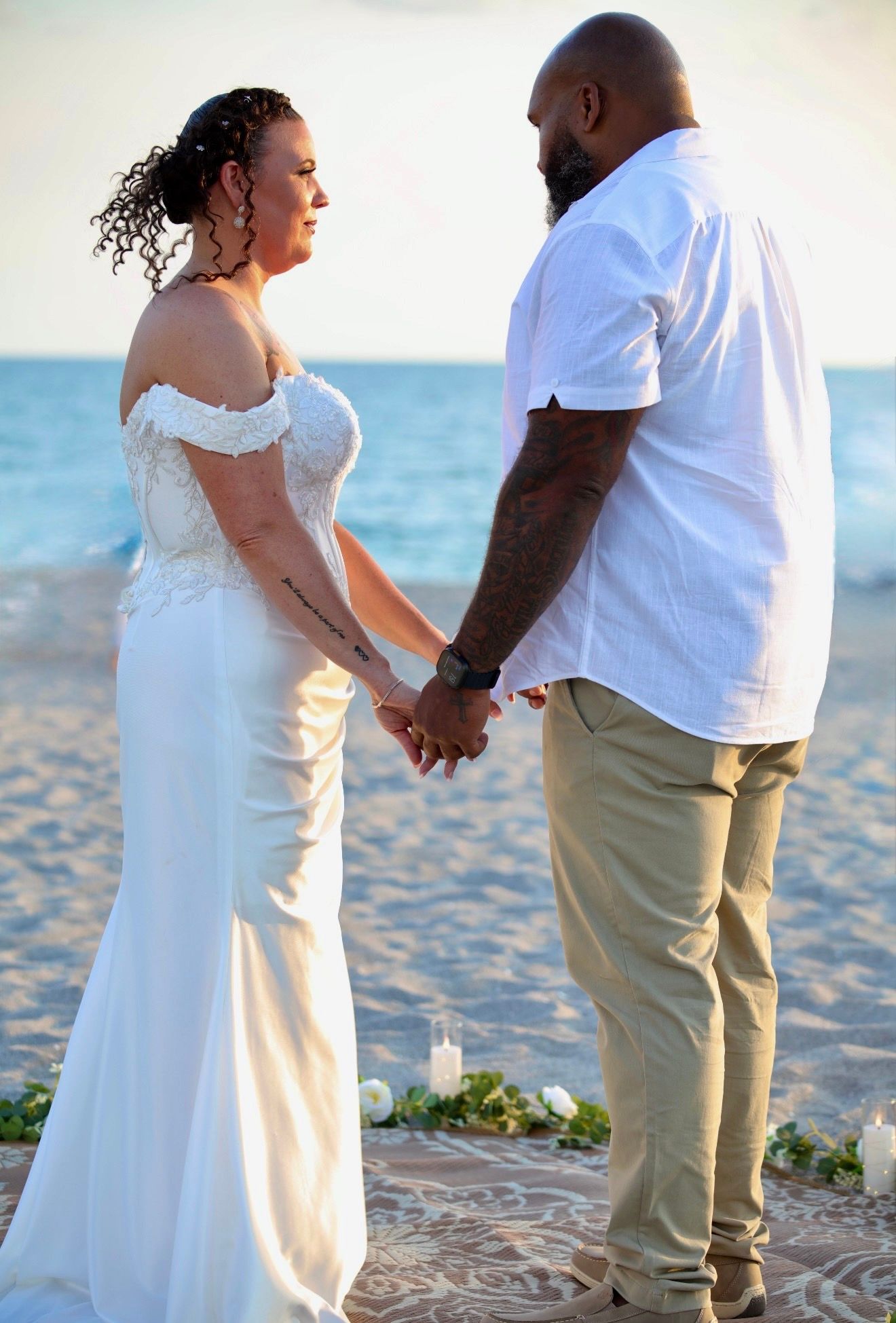 Couple exchanging vows on beach at sunset, bride in white off-shoulder gown holding grooms hands.
