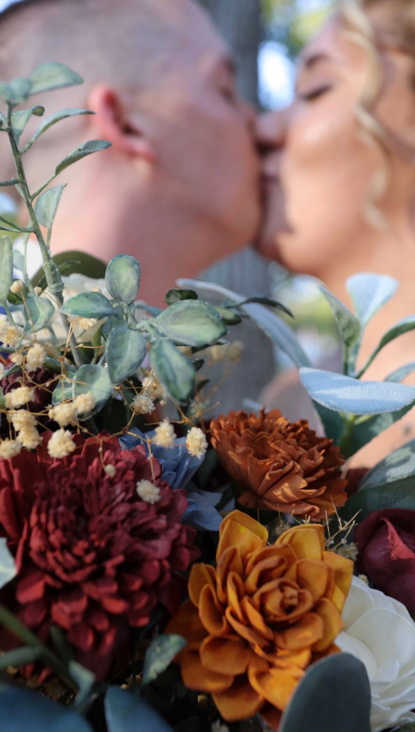Close-up of a colorful wedding bouquet with burgundy, rust, and gold flowers while bride and groom kiss softly in background.
