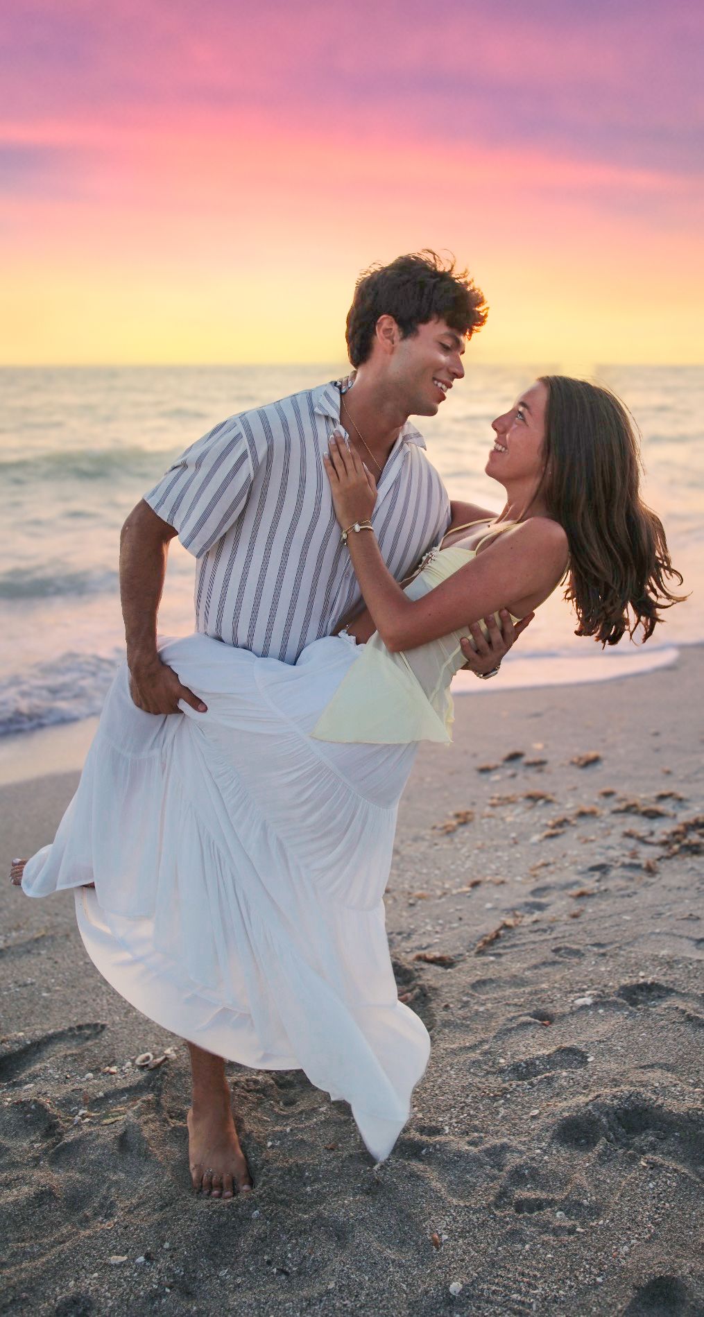 Couple dancing joyfully on beach at sunset, man lifting woman in white skirt and yellow top with colorful sky backdrop.
