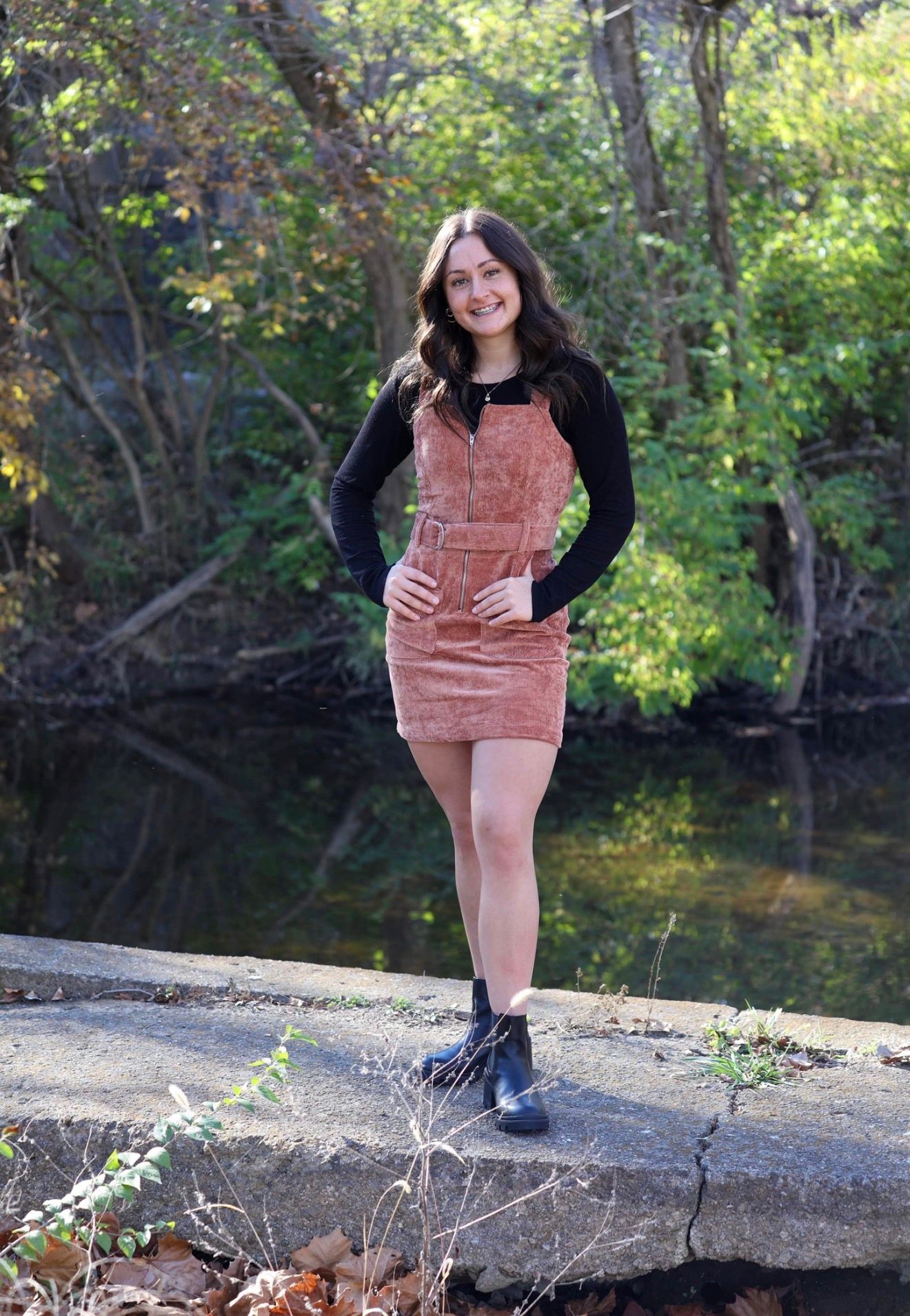 Young woman in pink corduroy overall dress and black boots smiling outdoors near a creek in autumn.