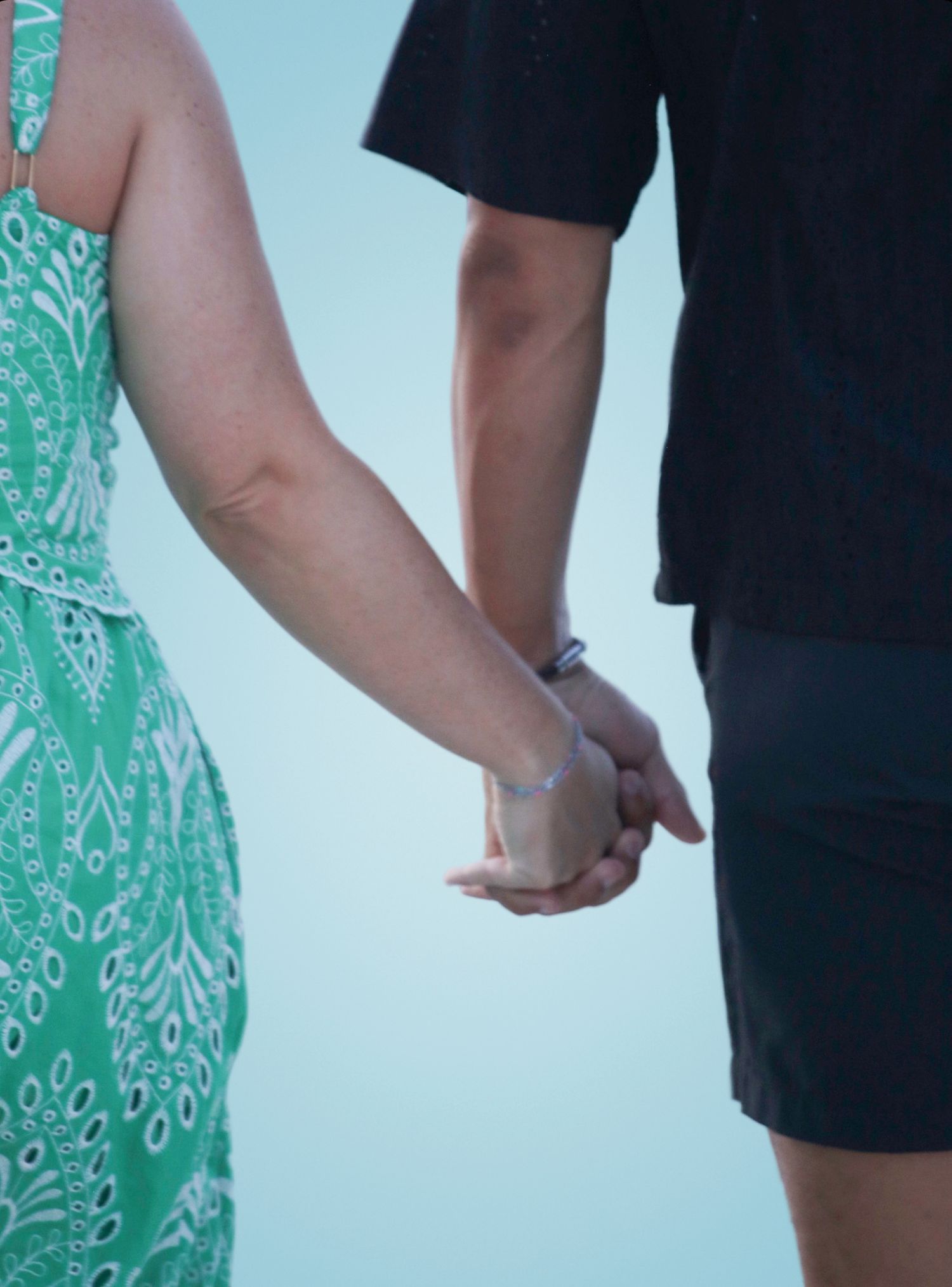 Couple holding hands at the beach, woman in green sundress and man in black shirt and shorts.