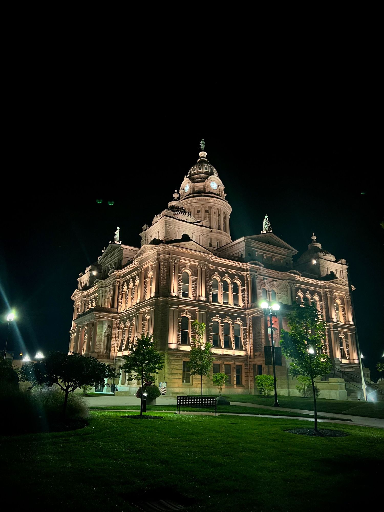 Historic courthouse illuminated at night with warm golden lights, lush green lawn, and ornate Victorian architecture under a dark sky.