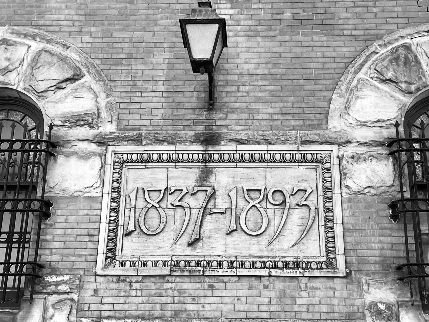 Black and white photo of a brick building facade with dates 1837-1893 carved in decorative stone plaque.