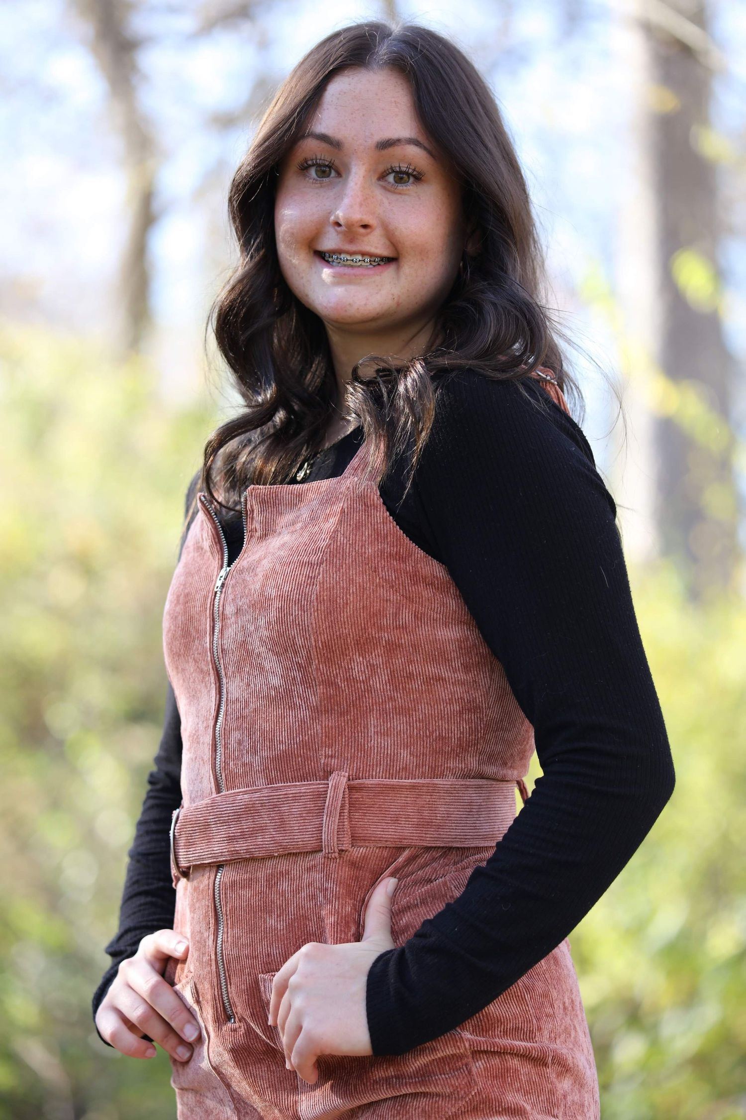 Young woman in pink corduroy overalls and black shirt smiling outdoors in a wooded setting.