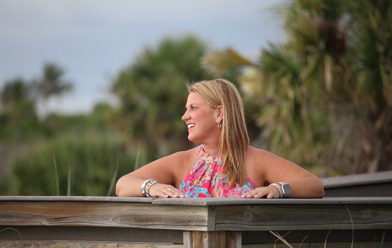 Smiling blonde woman in floral top leaning on wooden railing with tropical palm trees in background.