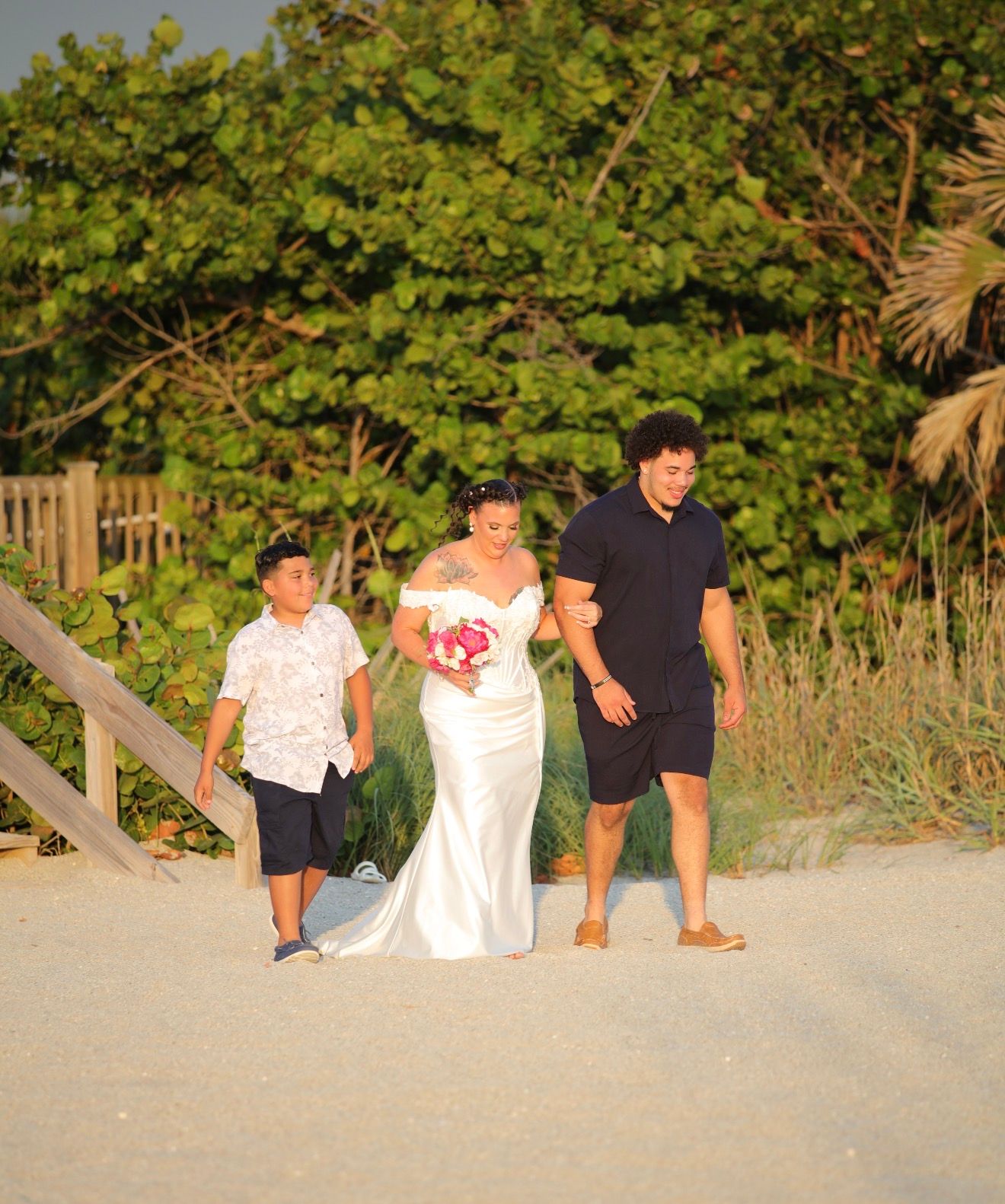 Bride in white gown holding pink bouquet walks on beach with two children during golden hour sunset.