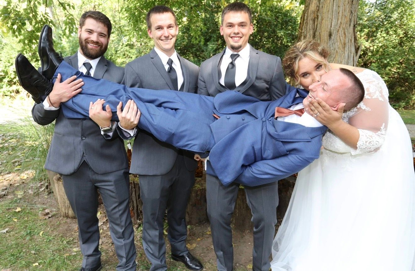 Wedding party photo with bride, groom carried by three groomsmen in gray suits outdoors near trees.