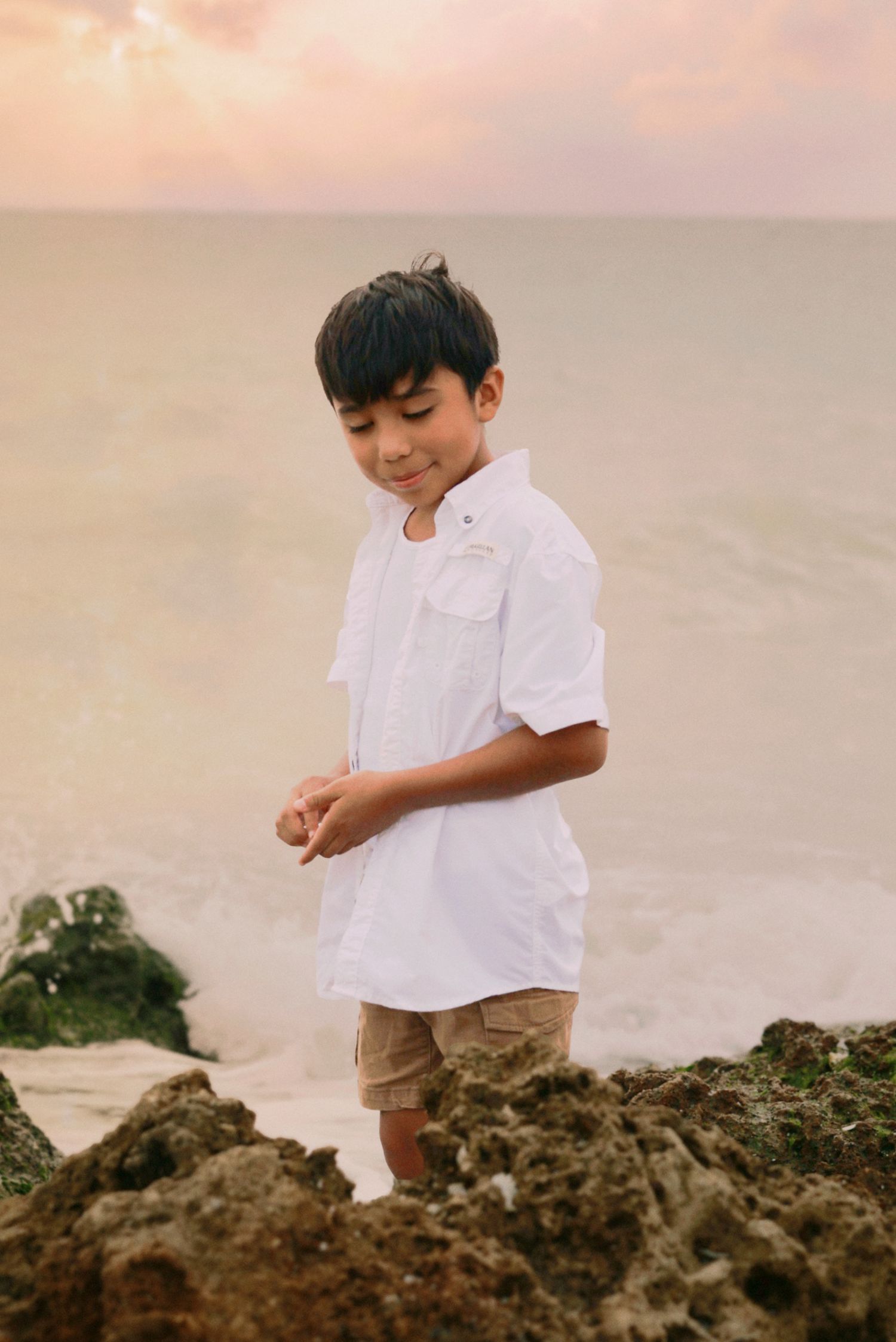 A young boy in a white shirt stands on rocky beach shore during a golden sunset.
