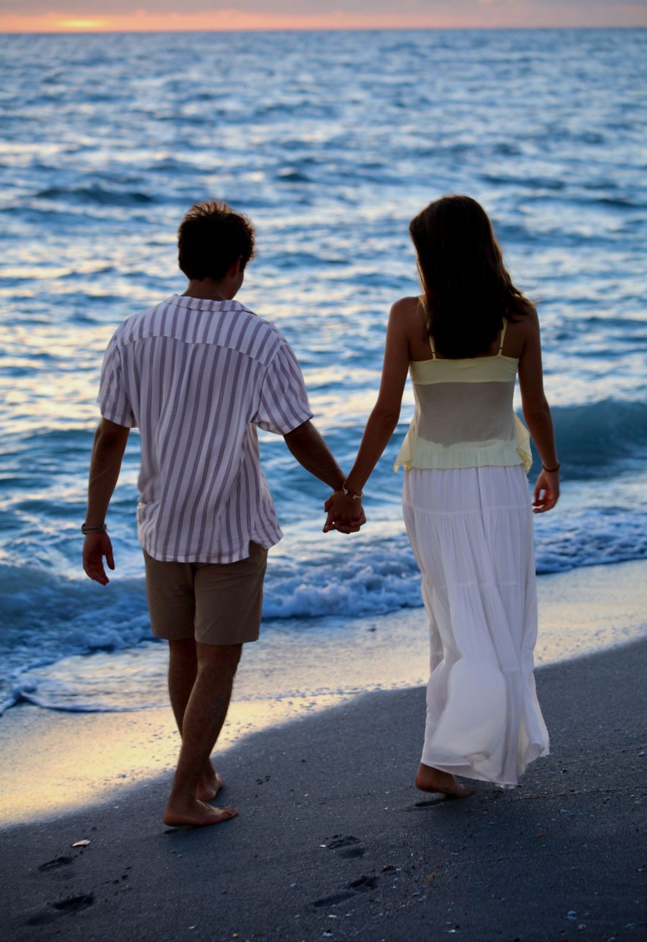 Romantic couple holding hands strolling along ocean shoreline at dusk, leaving footprints in the sand.