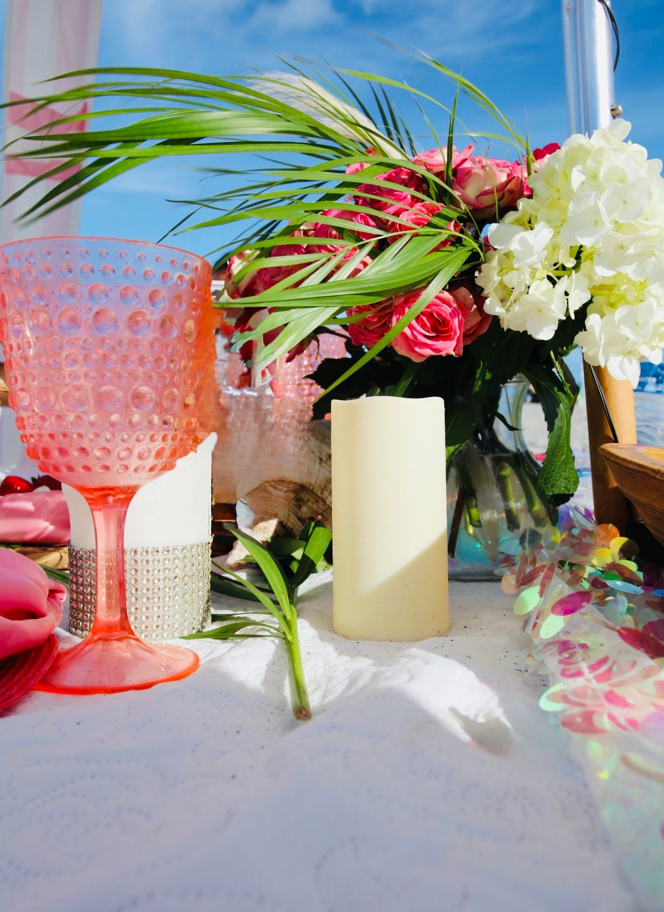 Tropical beach table with pink hobnail goblet, white hydrangeas, roses, palm leaves, and pillar candle.