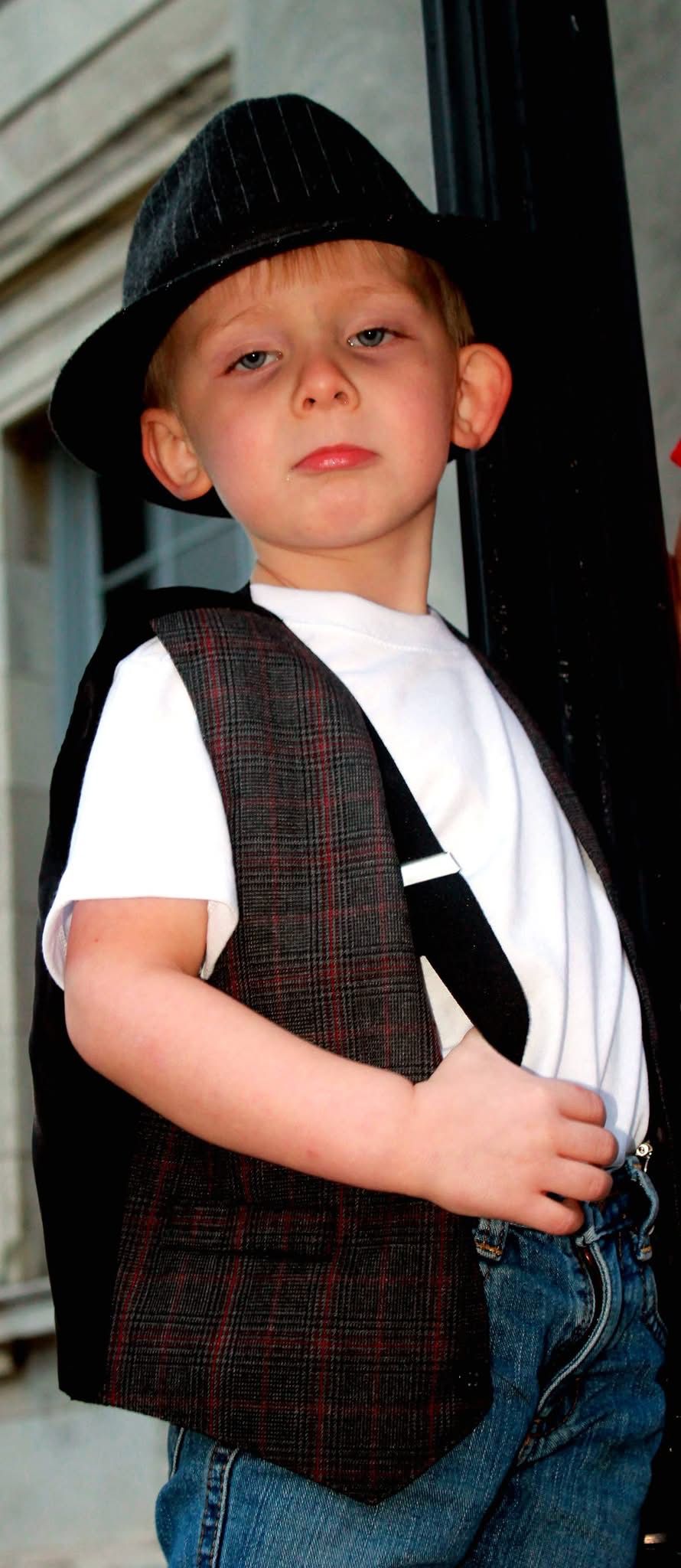 Young boy in a plaid vest, white shirt, jeans, and black fedora hat posing confidently outdoors.