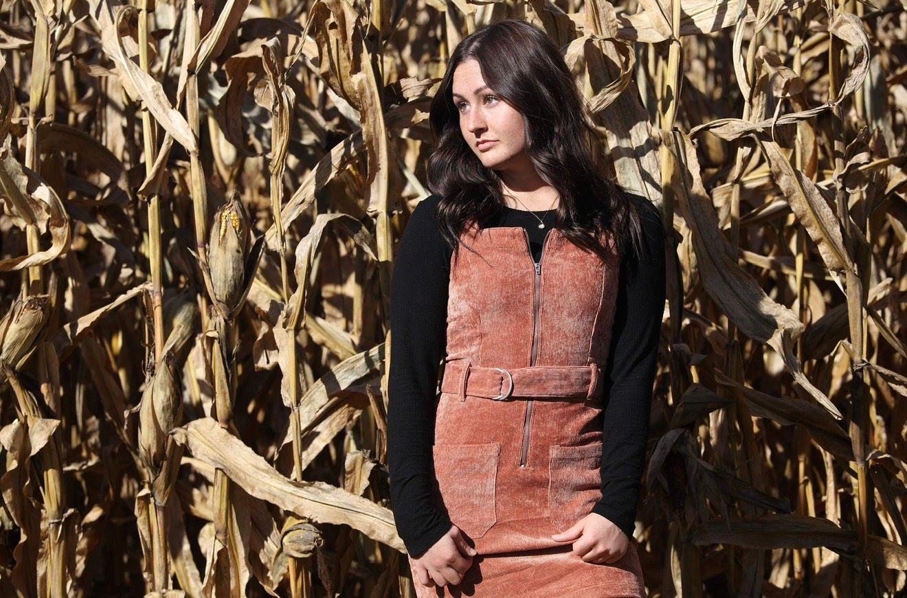 Woman in rust corduroy dress and black top posing in a dry autumn cornfield.