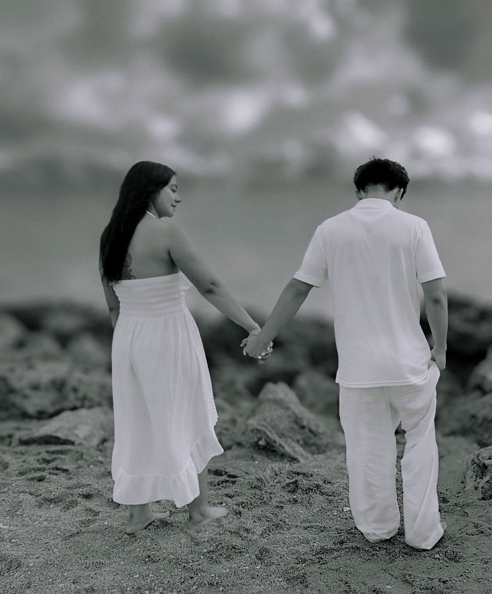 A couple in white clothing holds hands on a rocky beach under a cloudy sky in this black and white photo.