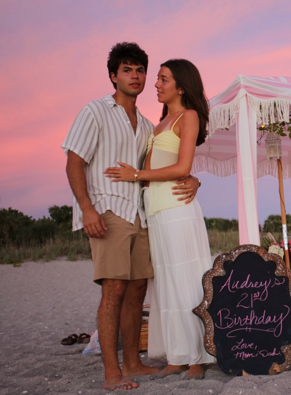 Couple embracing on beach at sunset beside Audrey's 21st Birthday sign with pink sky backdrop. Paradise Picnics in Venice, FL.