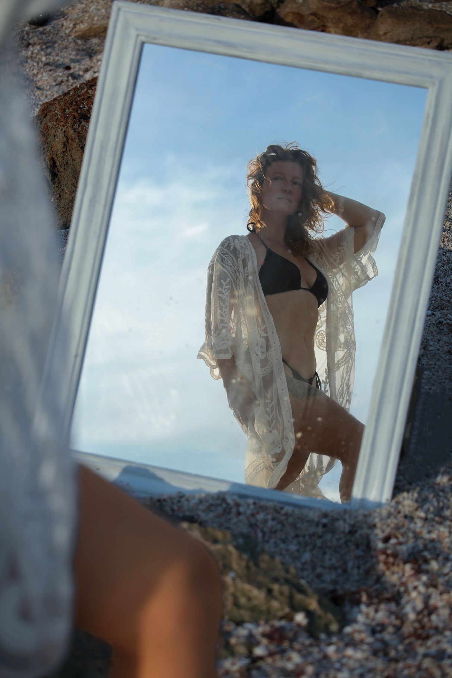 Woman in black bikini and white lace cover-up reflected in a vintage white frame mirror propped on sandy beach rocks.