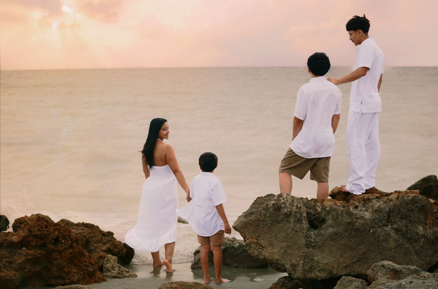 Family in white clothing standing on rocks by the ocean at sunset, enjoying a serene beach view together.