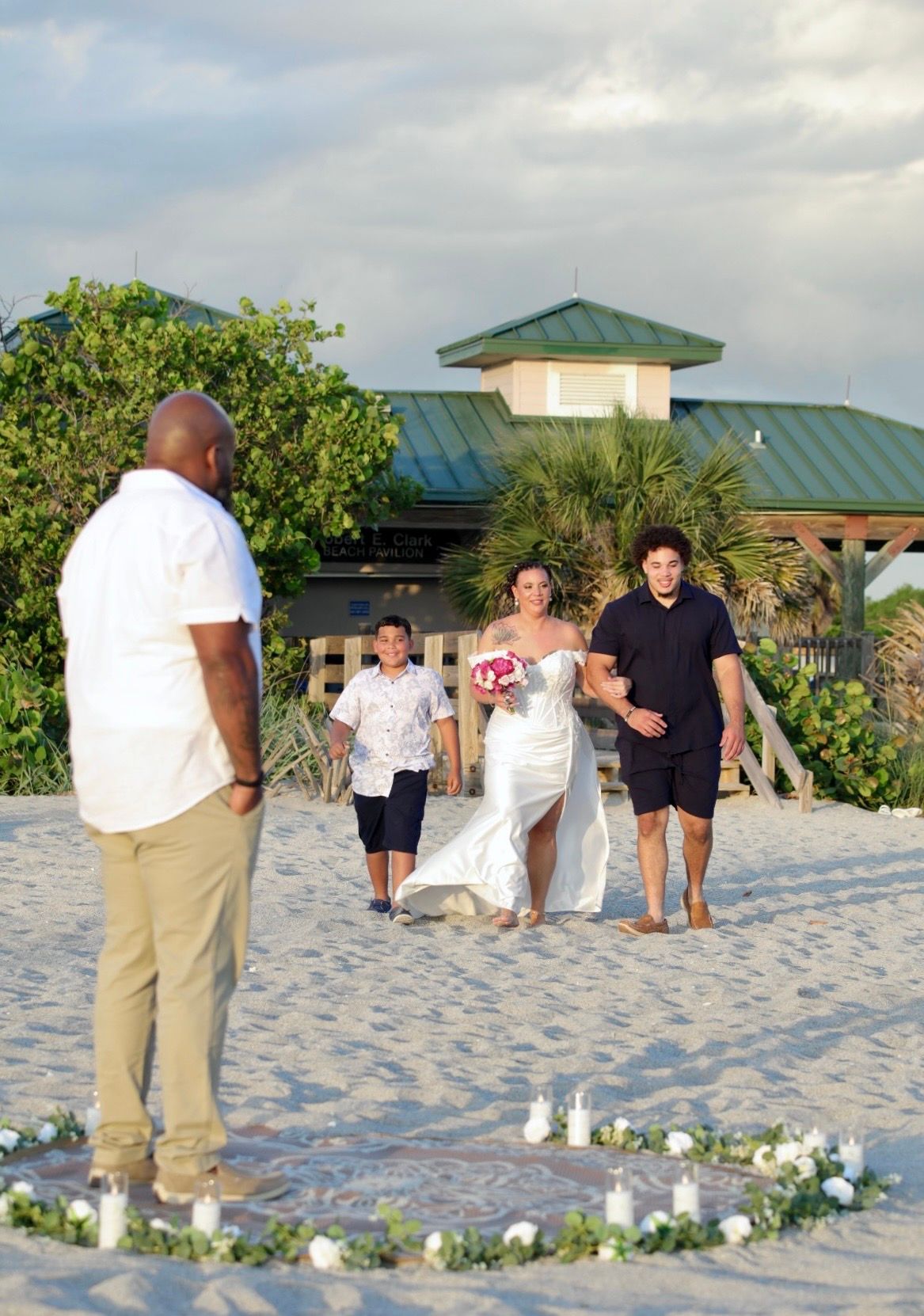Bride walks down sandy beach aisle with two boys toward groom, candles and floral greenery marking the ceremony space.