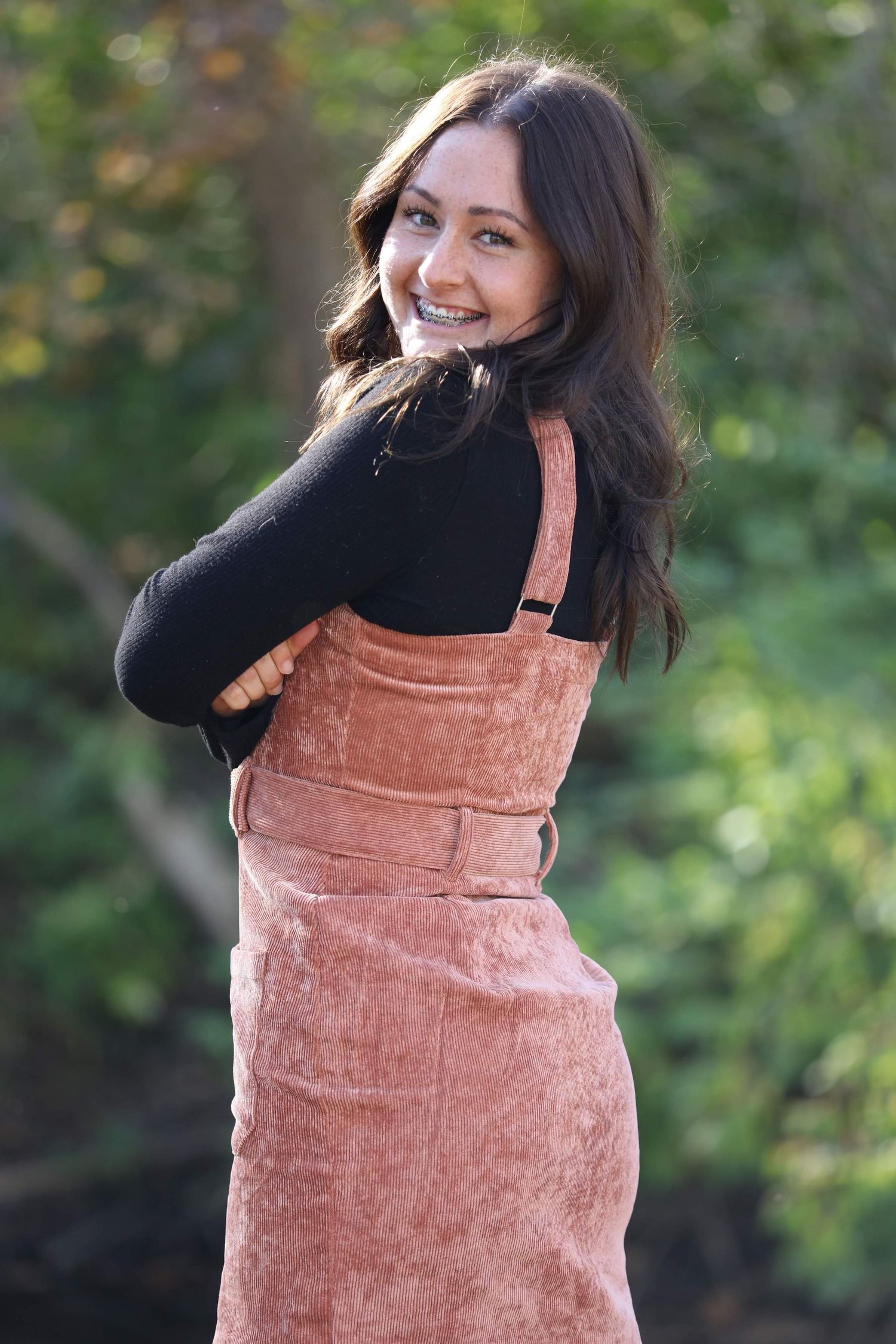 Smiling brunette teen in pink corduroy overalls and black top posing outdoors with arms crossed.
