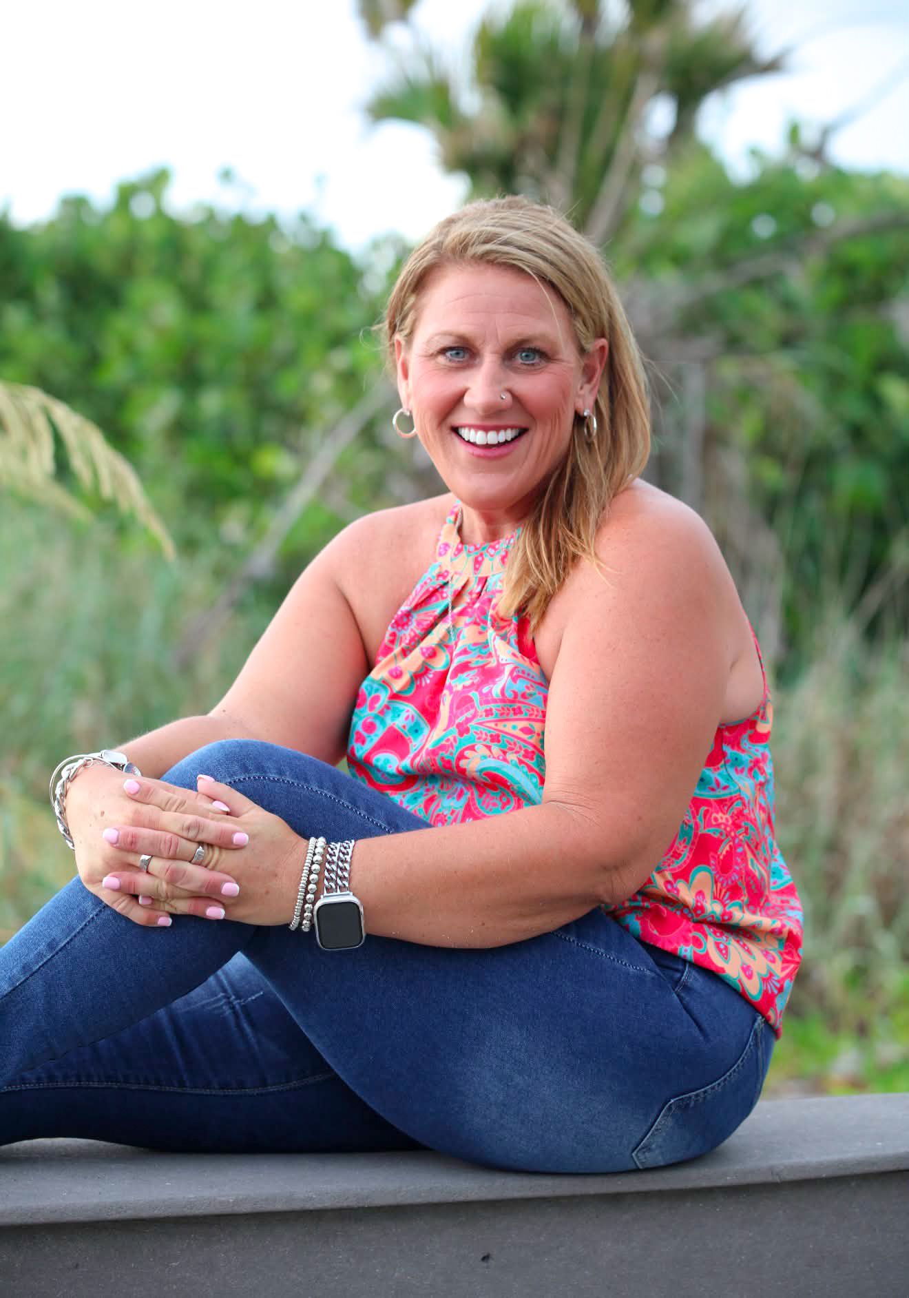 Smiling blonde woman in colorful floral top and jeans sitting outdoors with lush green foliage background.