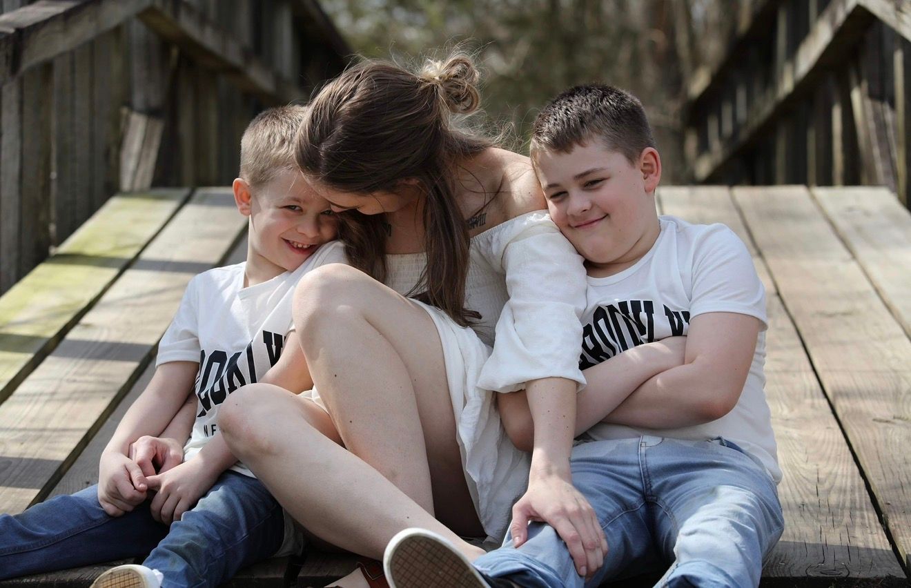 A mother sits on a wooden bridge hugging her two young sons, all wearing matching white graphic tees.