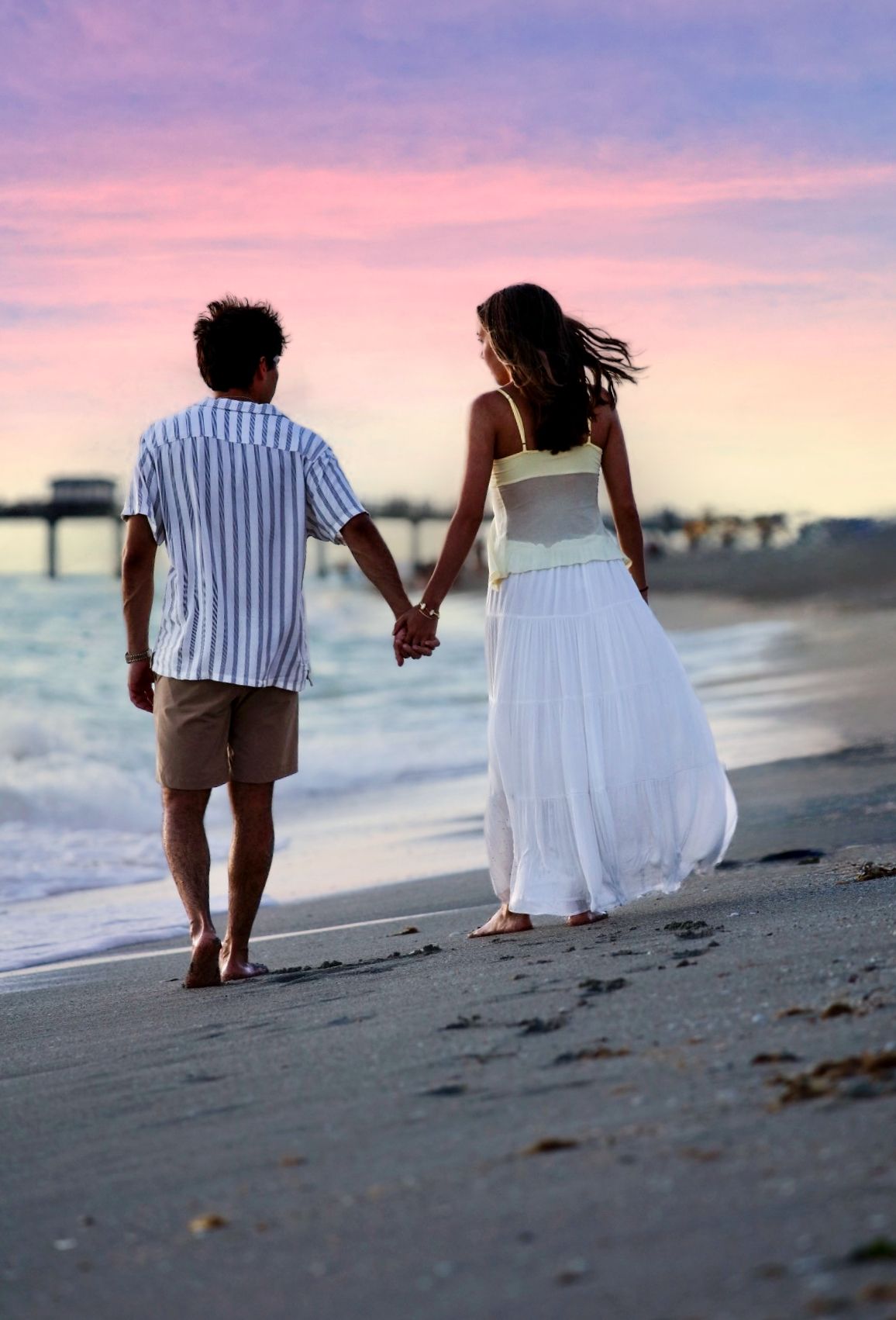 Couple holding hands walking barefoot on beach at colorful sunset, man in striped shirt and woman in white dress.