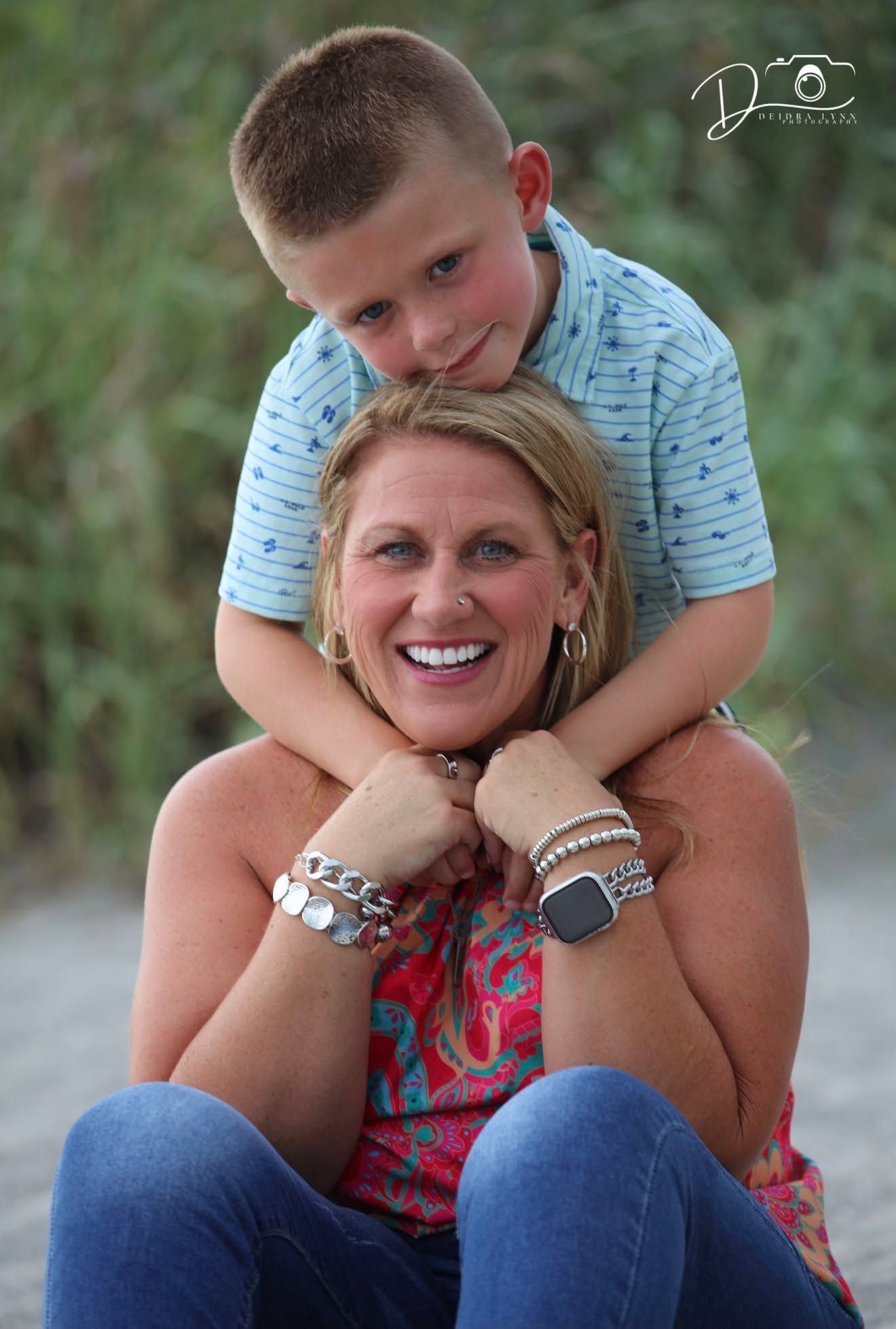 A smiling woman in a colorful top sits as a young boy in a blue striped shirt leans over her shoulders, posing for a portrait.