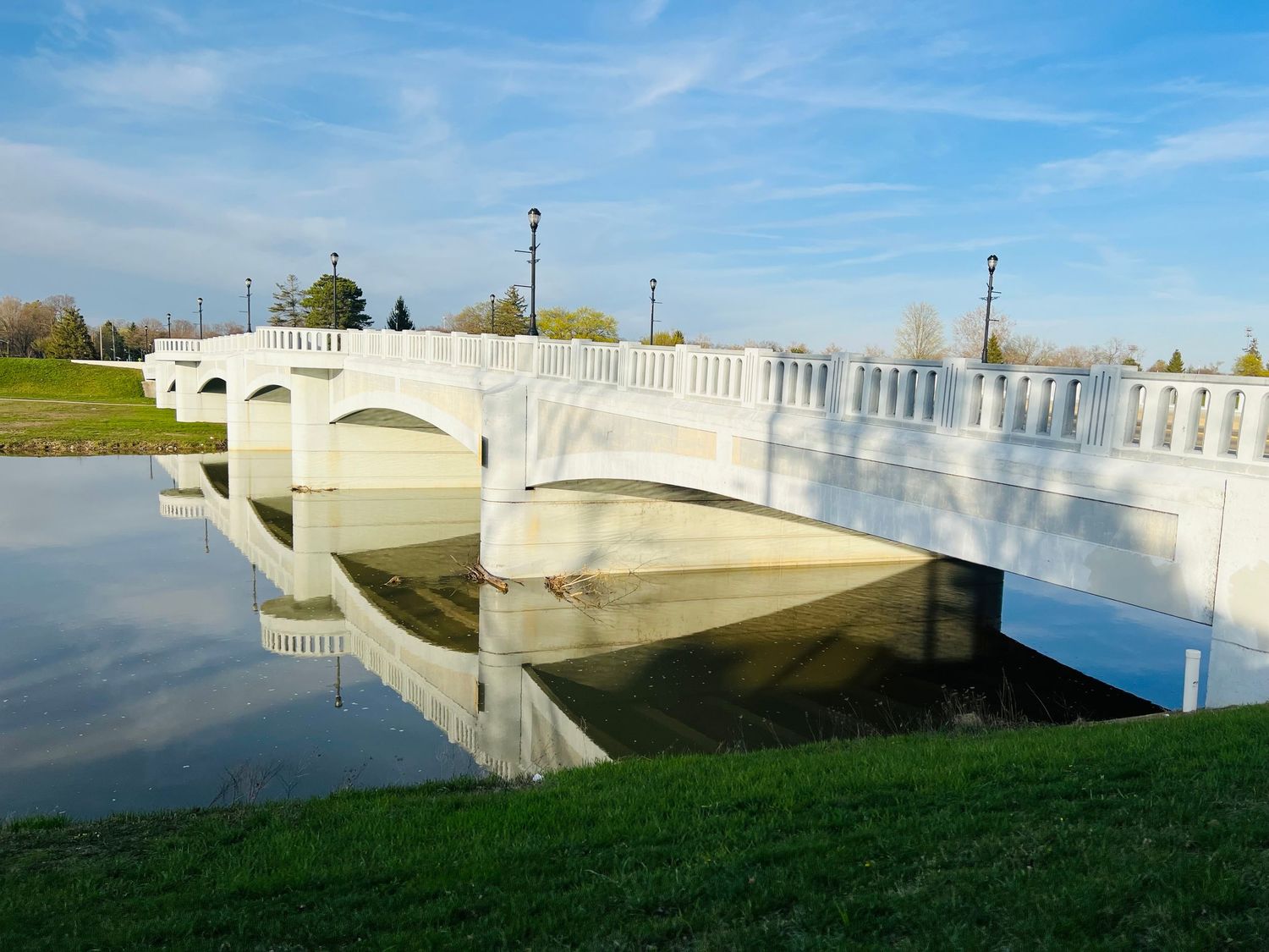 White arched bridge with decorative railings reflects in calm water, surrounded by green grass and trees under a blue sky.