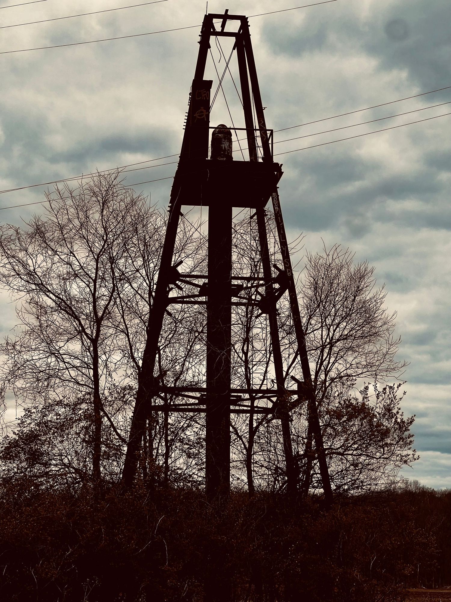 Silhouette of an old rustic oil derrick tower against a cloudy sky, surrounded by bare trees in a rural landscape.