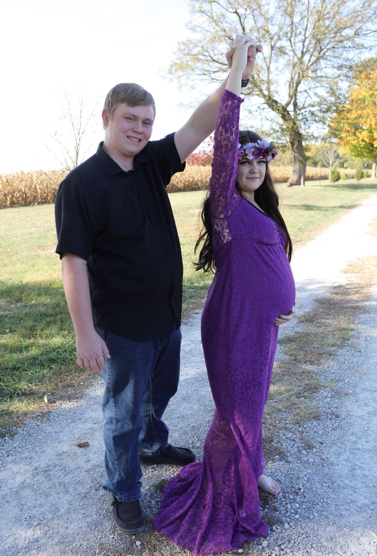 Pregnant woman in purple lace maternity gown held by partner on rural path during fall outdoor maternity photoshoot.