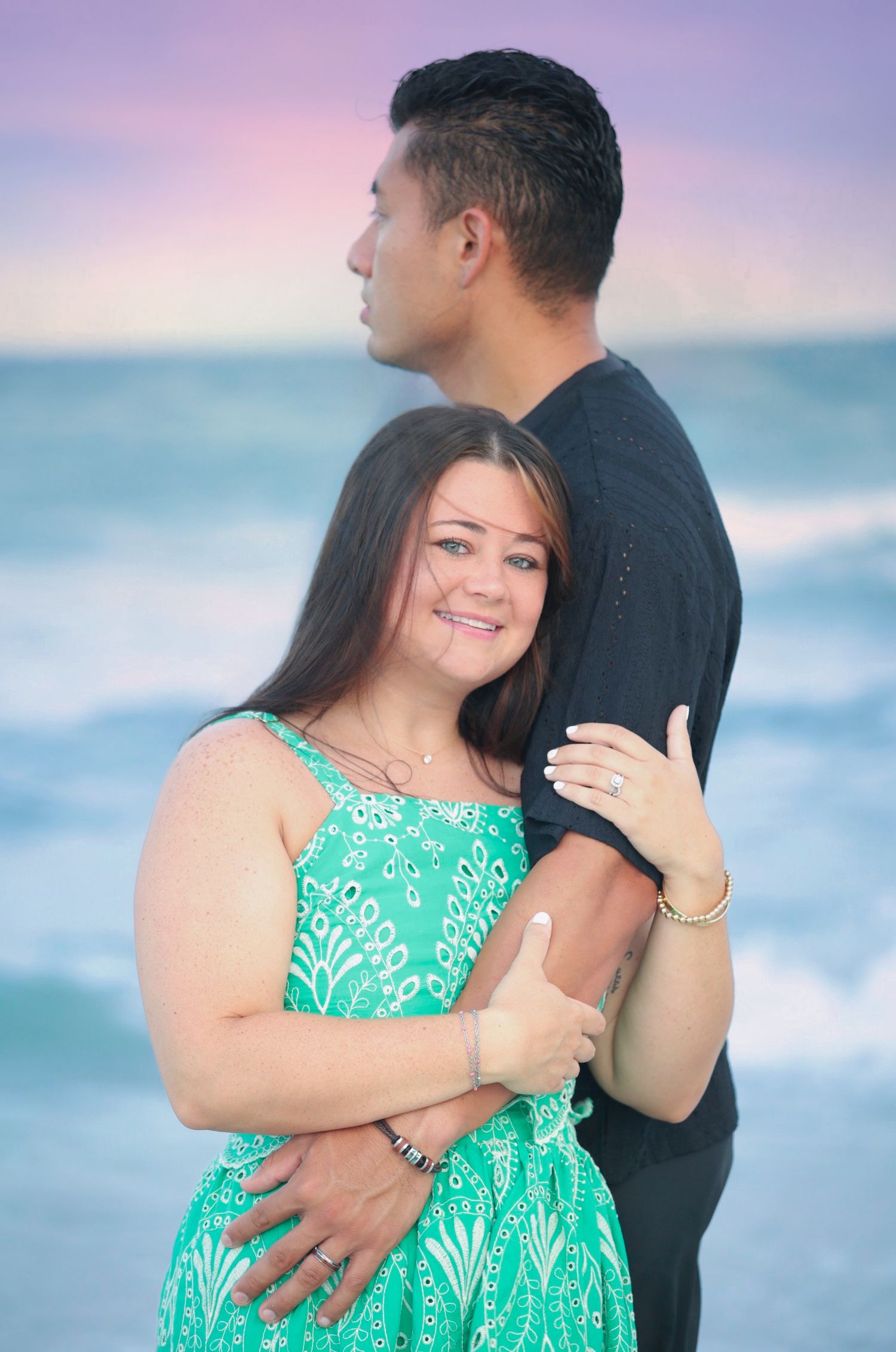 Smiling woman in teal dress embraced by man in black shirt on a beach at sunset, ocean in background.