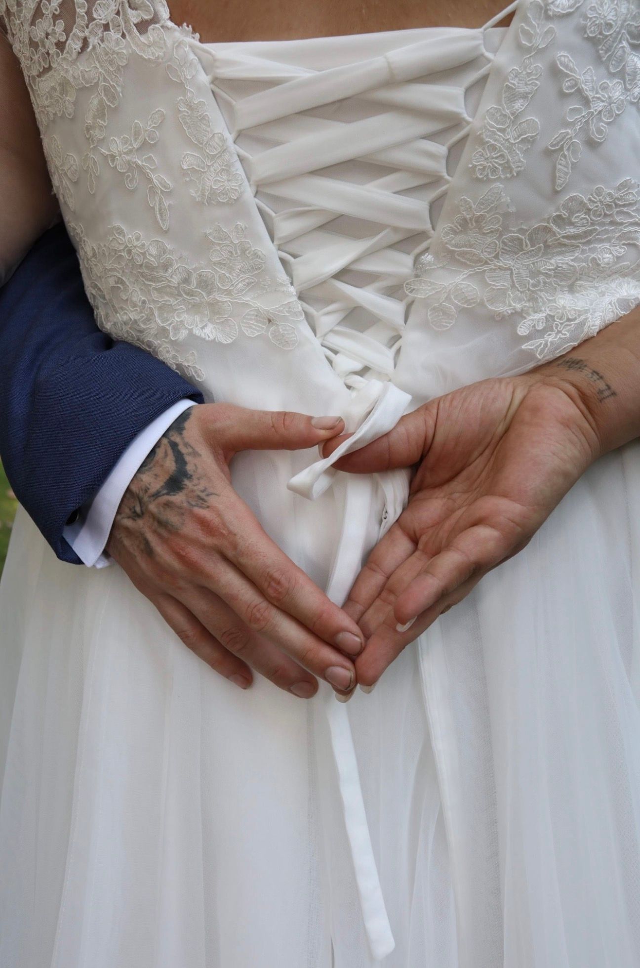 Bride and groom holding hands at the back of a lace wedding dress with corset ribbon detail, tattoos visible.