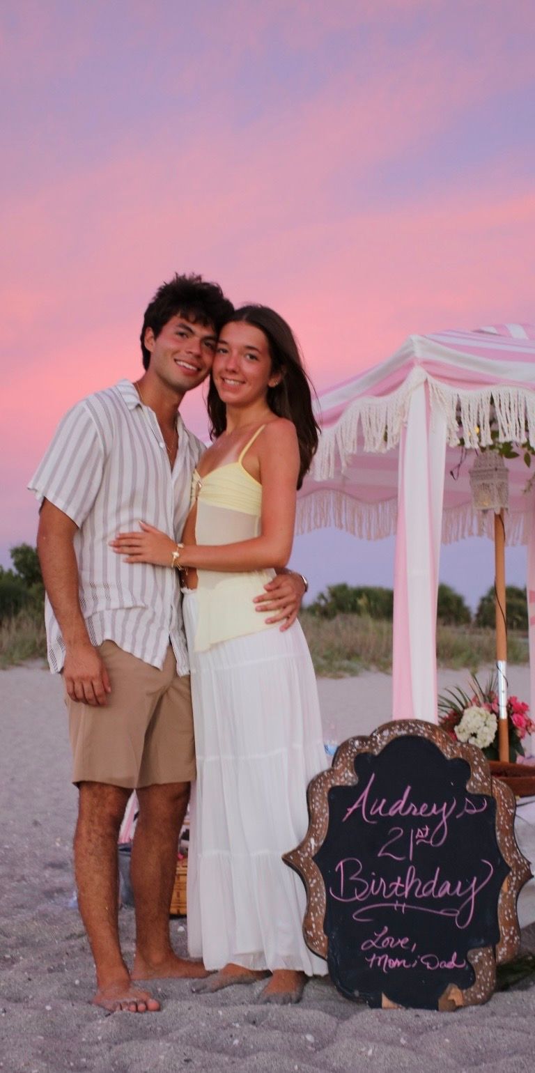Couple embracing on beach at sunset beside Audrey's 21st Birthday sign from Mom and Dad.