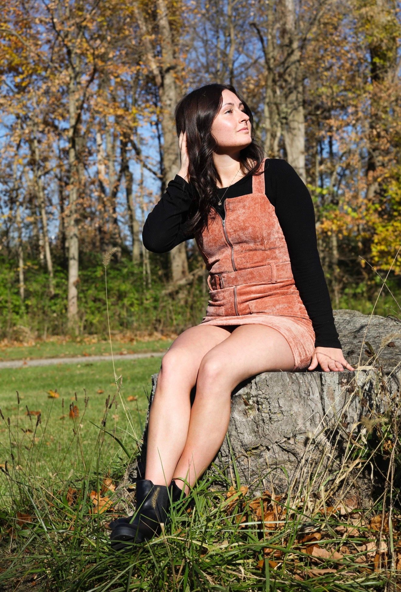 Young woman in rust overalls and black turtleneck sits on a rock in an autumn park, gazing upward.