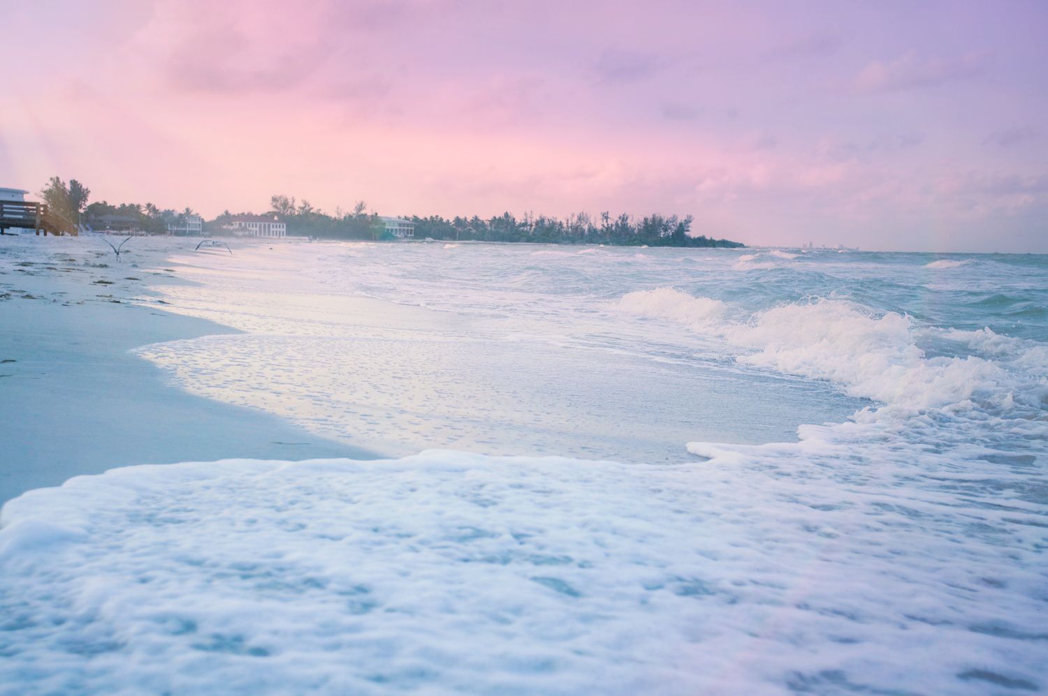 Pastel pink and purple sunset sky over a tropical beach with white foamy waves washing onto white sand shores.