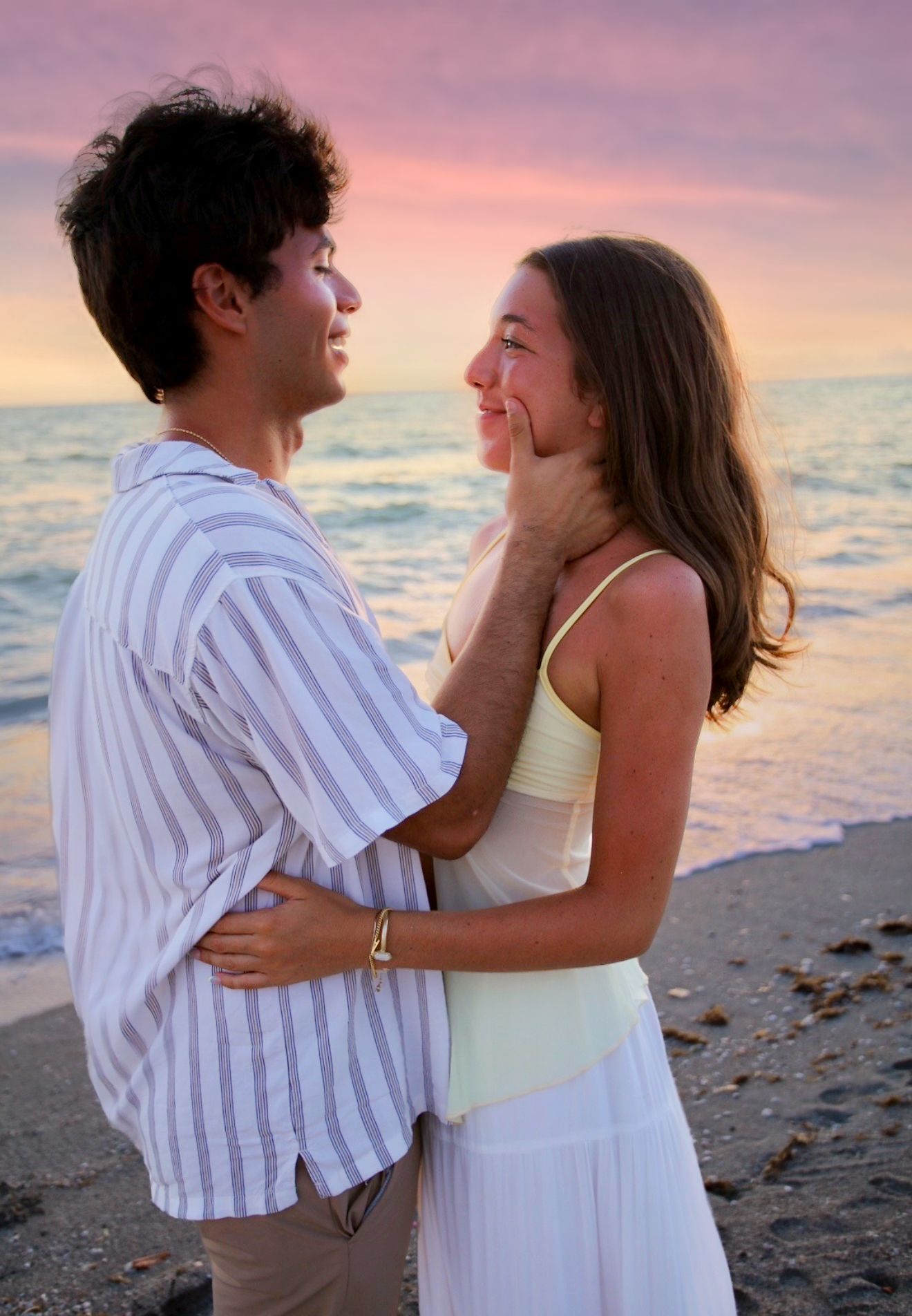 A smiling young couple embraces on a sandy beach during a colorful pink and purple sunset.