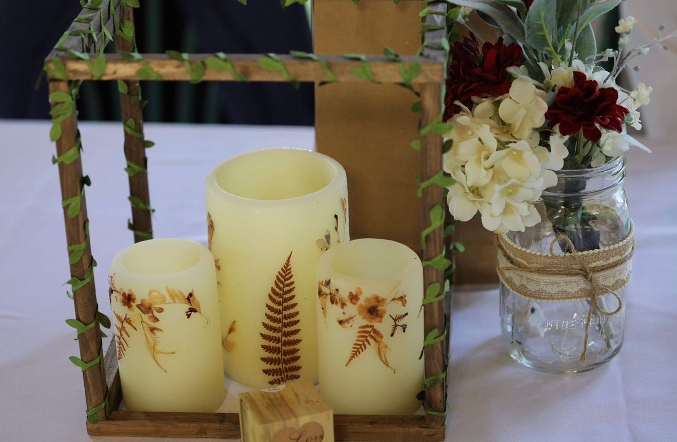 Three cream pillar candles with fern prints in a wooden frame beside a mason jar with red and white flowers.