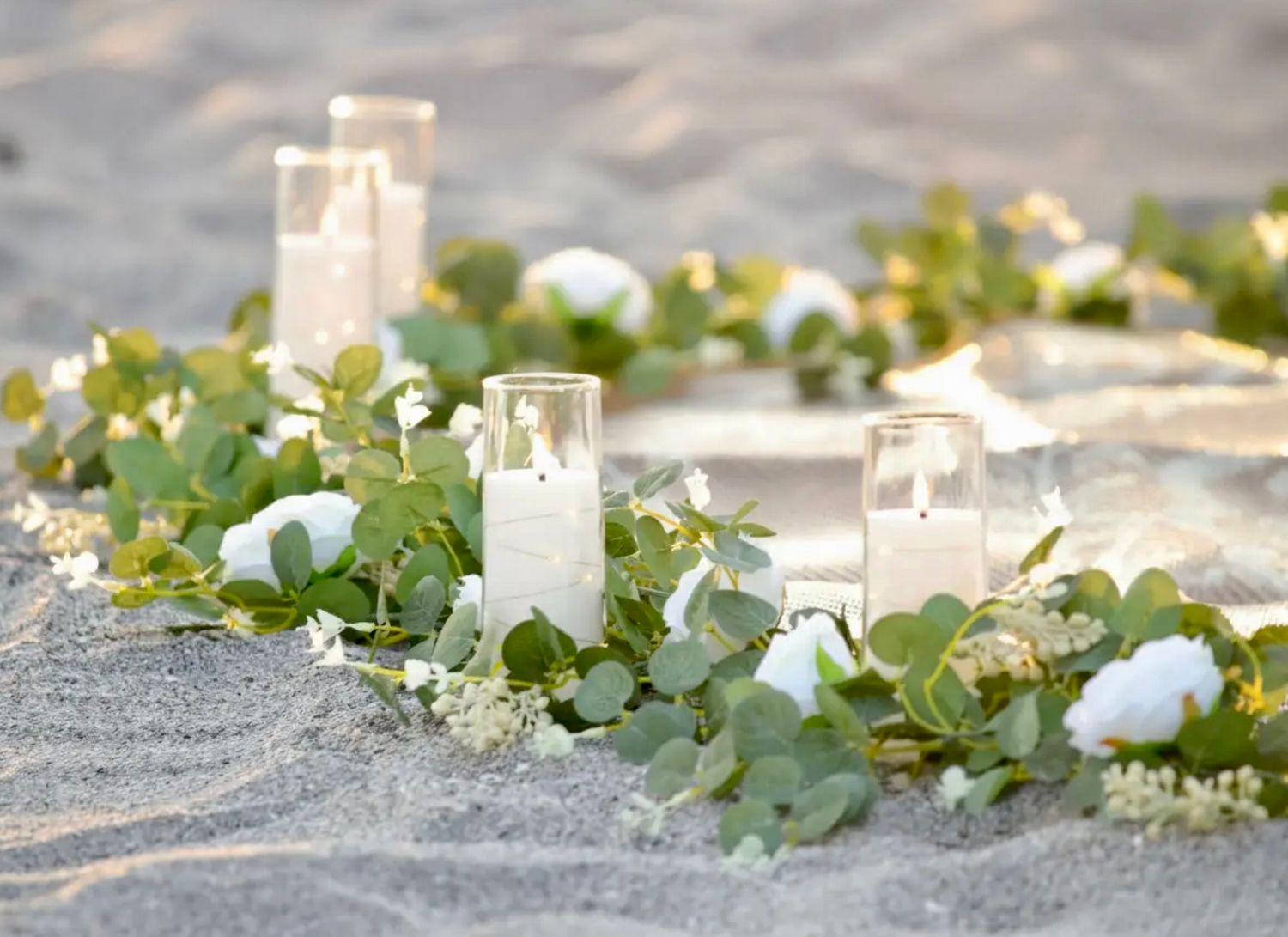 White candles and eucalyptus garland with white roses arranged on sandy beach for elegant outdoor wedding ceremony.
